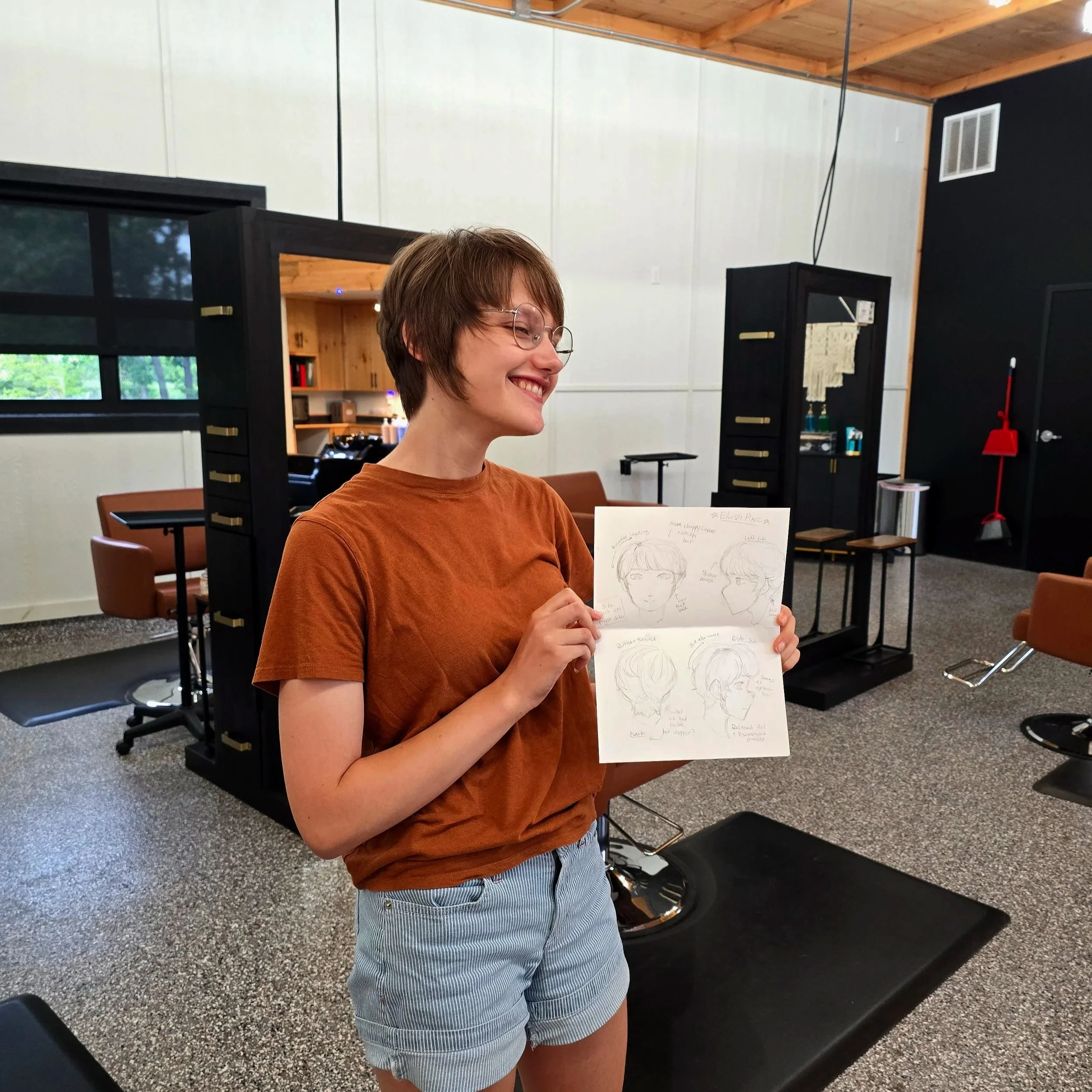 A young woman with short brown hair, glasses, and a brown t-shirt, smiling and holding a sketchbook with various character drawings inside a hair salon or barbershop.