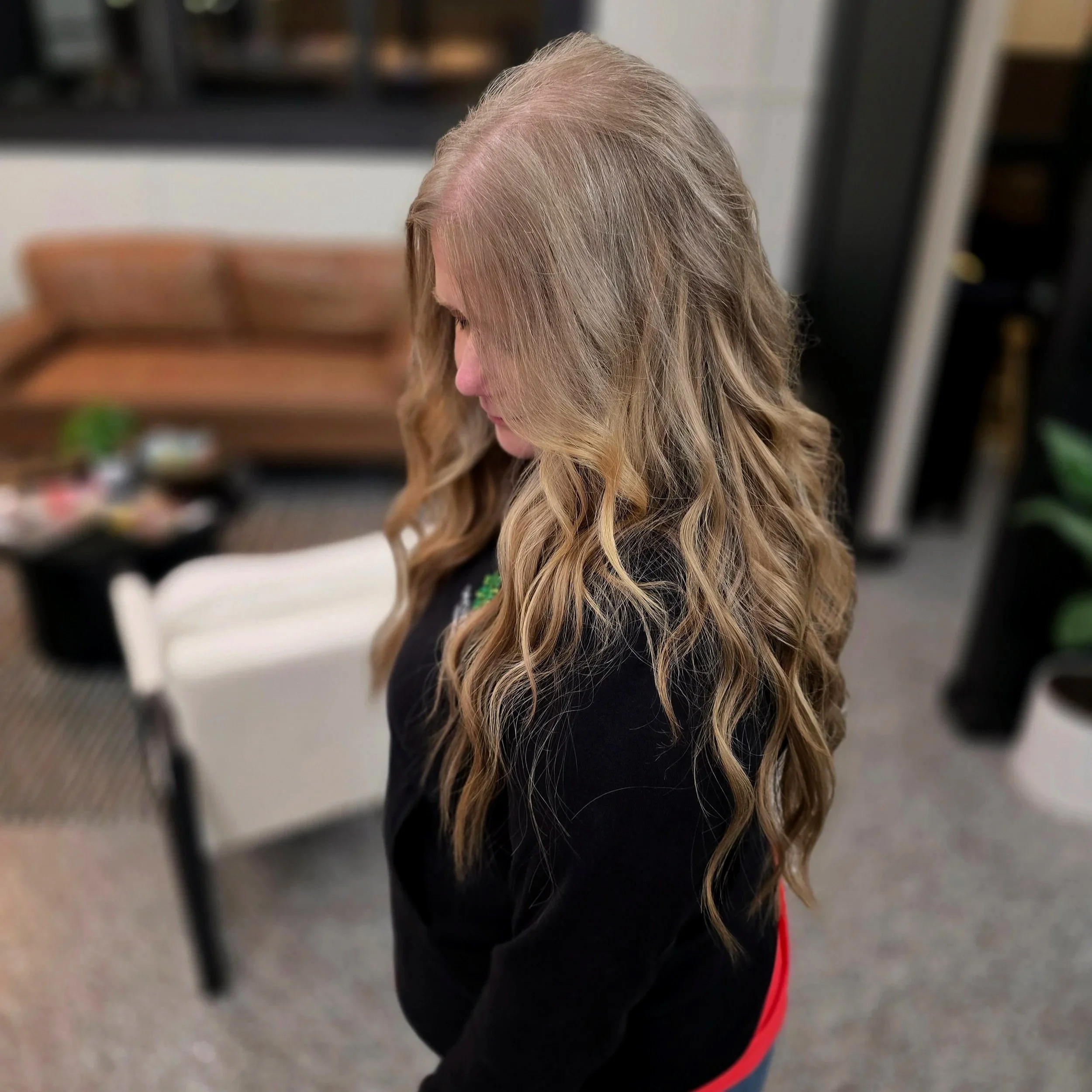 A woman with long, wavy blonde hair looking downward indoors, with a blurred background of furniture and plants.