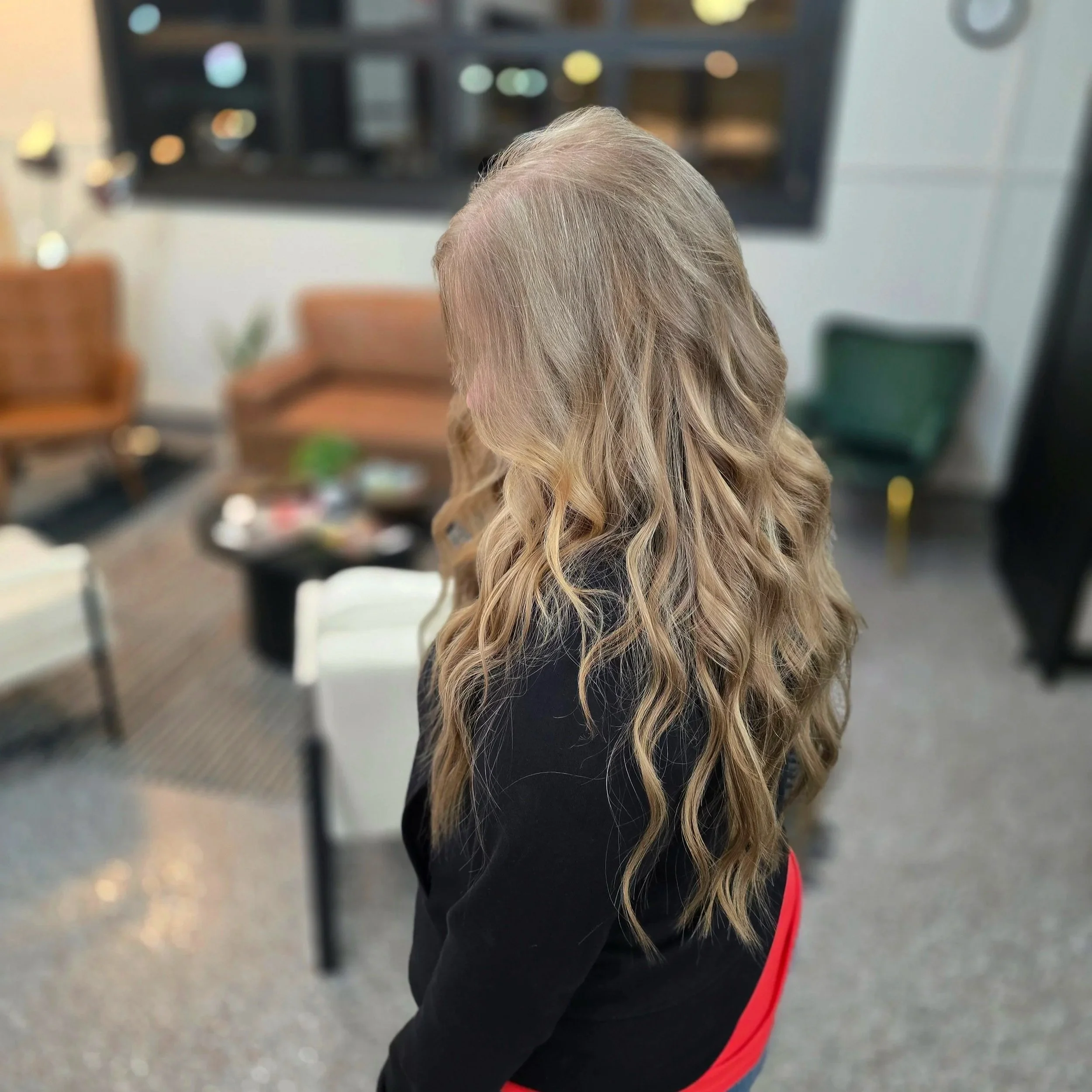 A woman with long, wavy blonde hair stands indoors in a modern room with colorful chairs and a coffee table in the background.