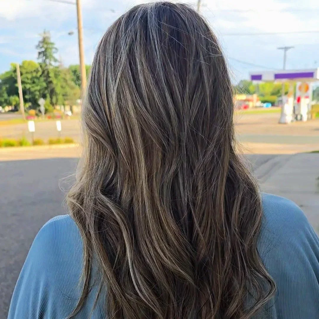 Back of a woman with long, wavy, dark hair standing outdoors, with trees, a road, and some buildings in the background.