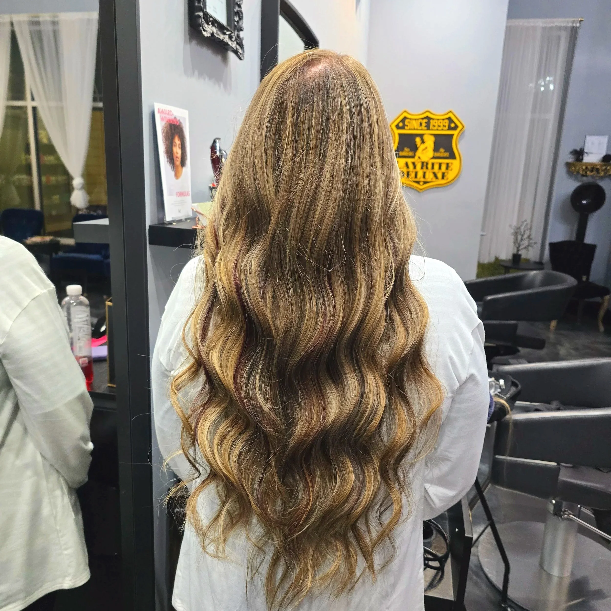 Back view of a woman with long, wavy blonde hair in a hair salon.