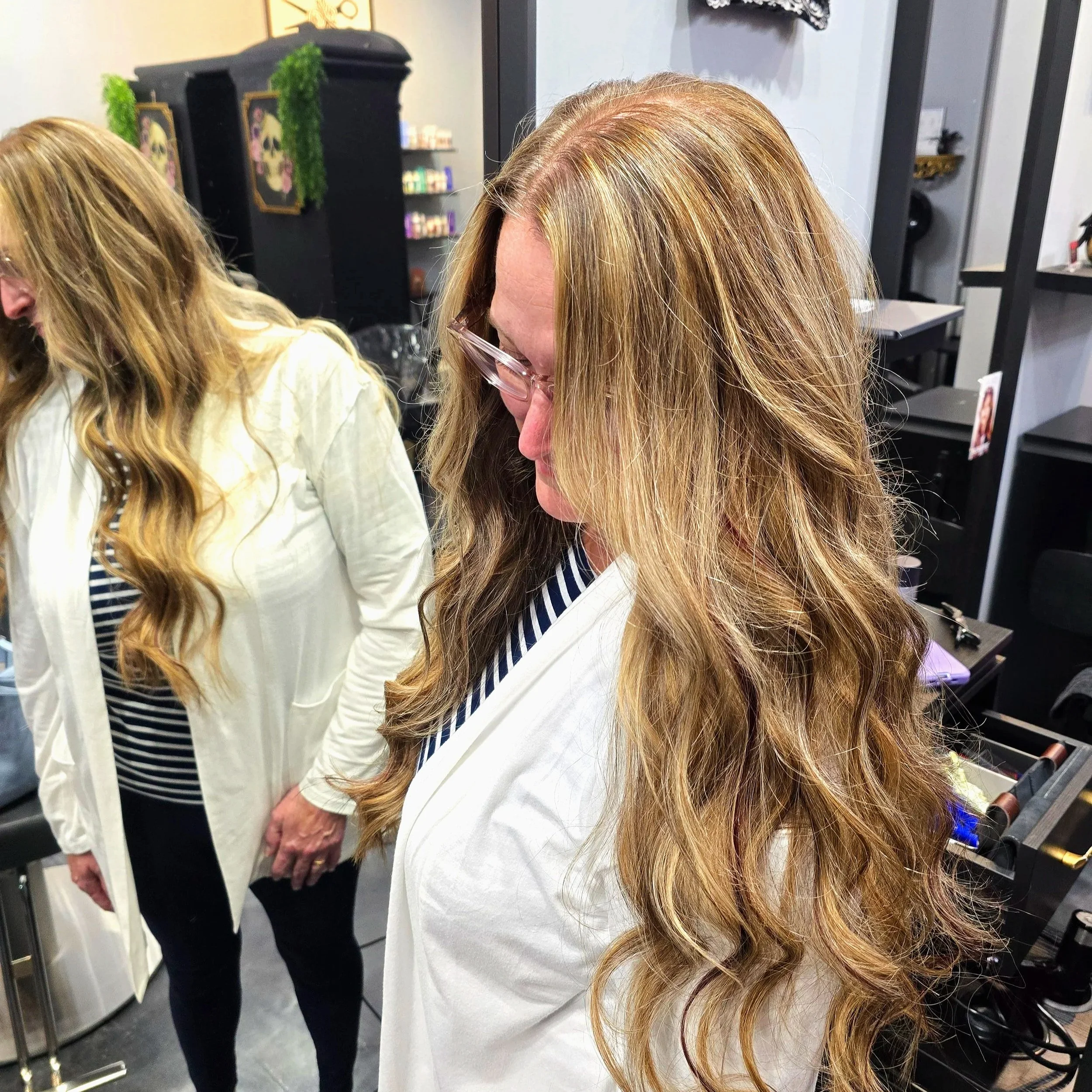 Woman with long, wavy, blonde hair wearing a white coat and glasses, standing in a hair salon.