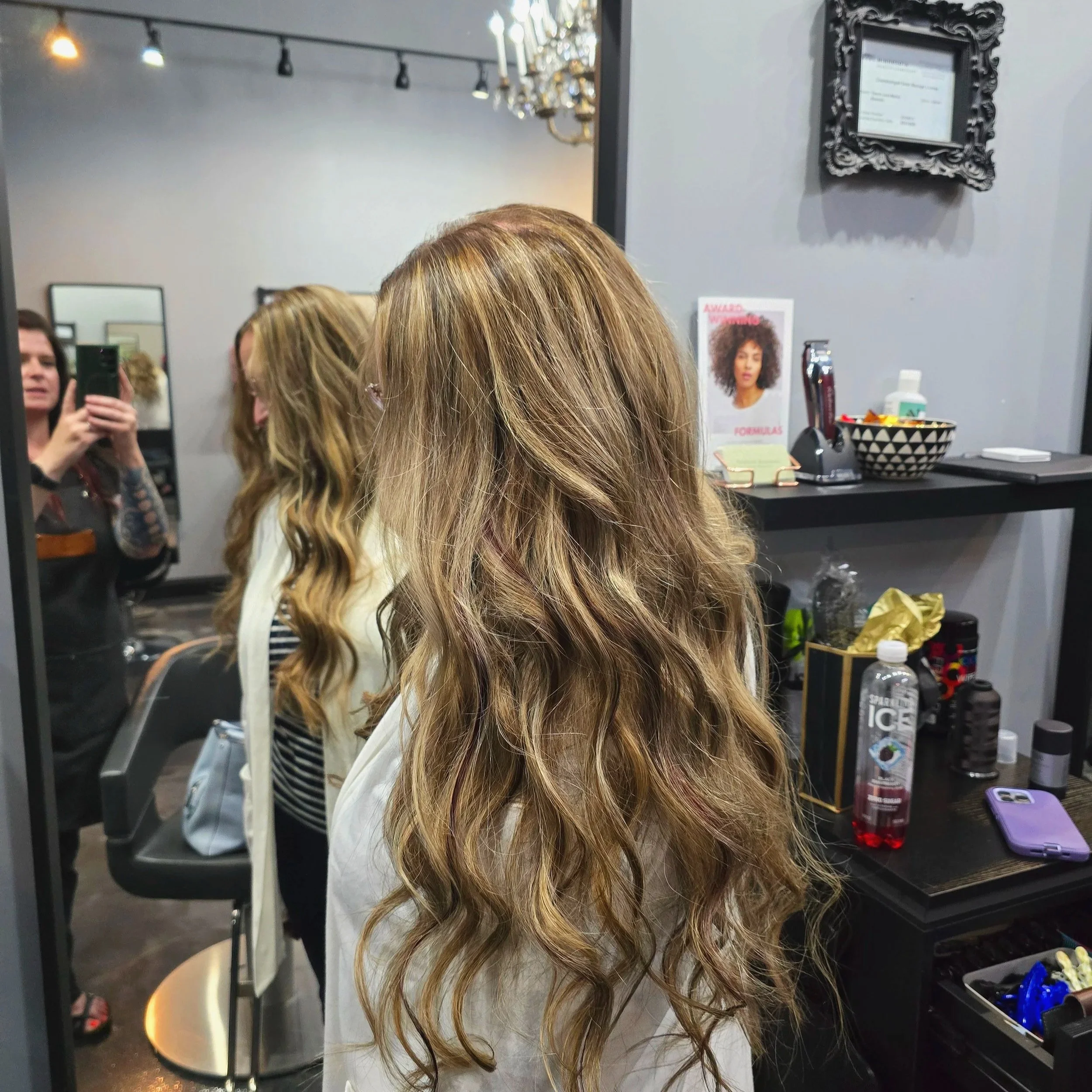 Woman with long, wavy, caramel-colored hair sitting in a hair salon with others getting hair services, a mirror, and salon products visible.