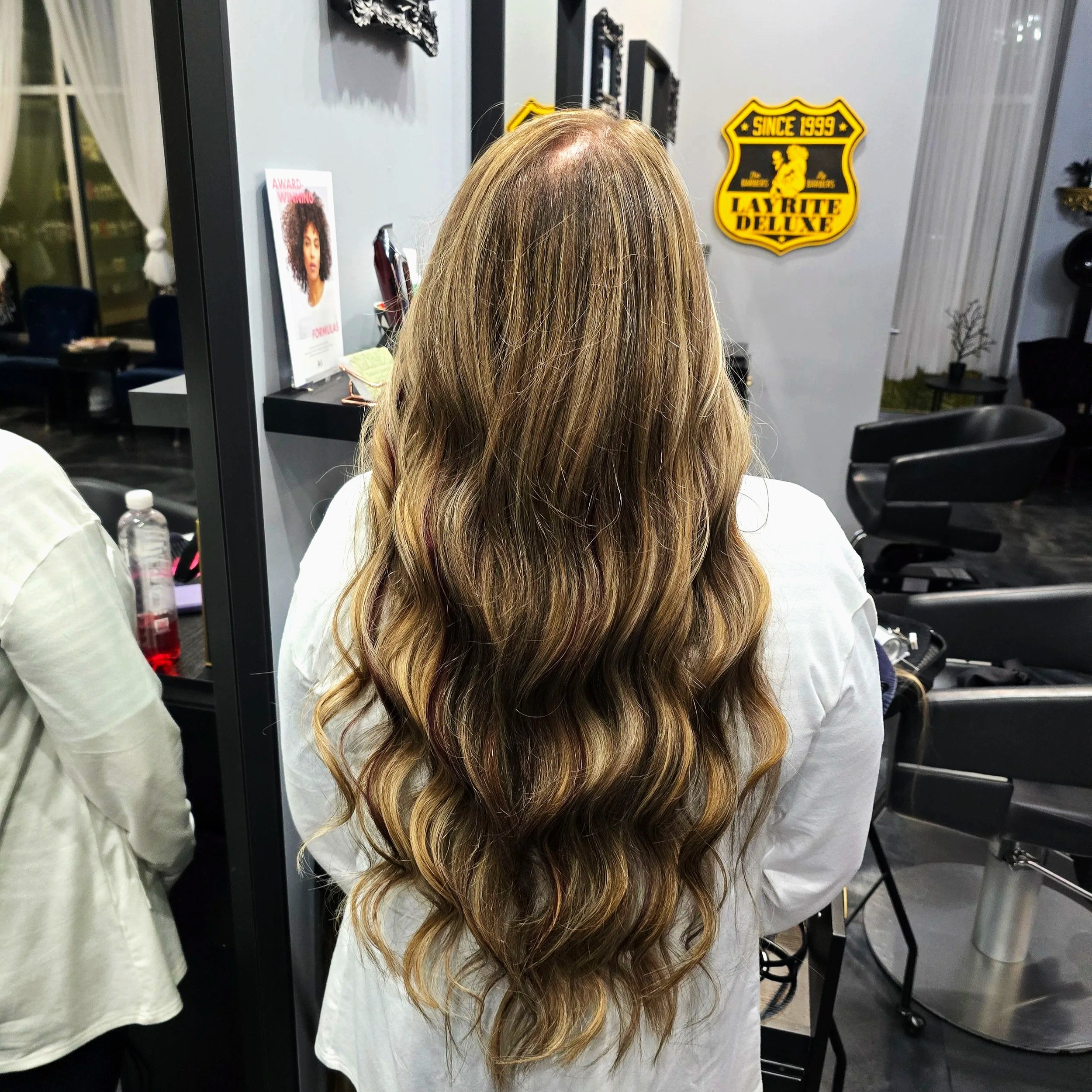 Back view of a woman with long, wavy, light brown hair with highlights, sitting in a hair salon.