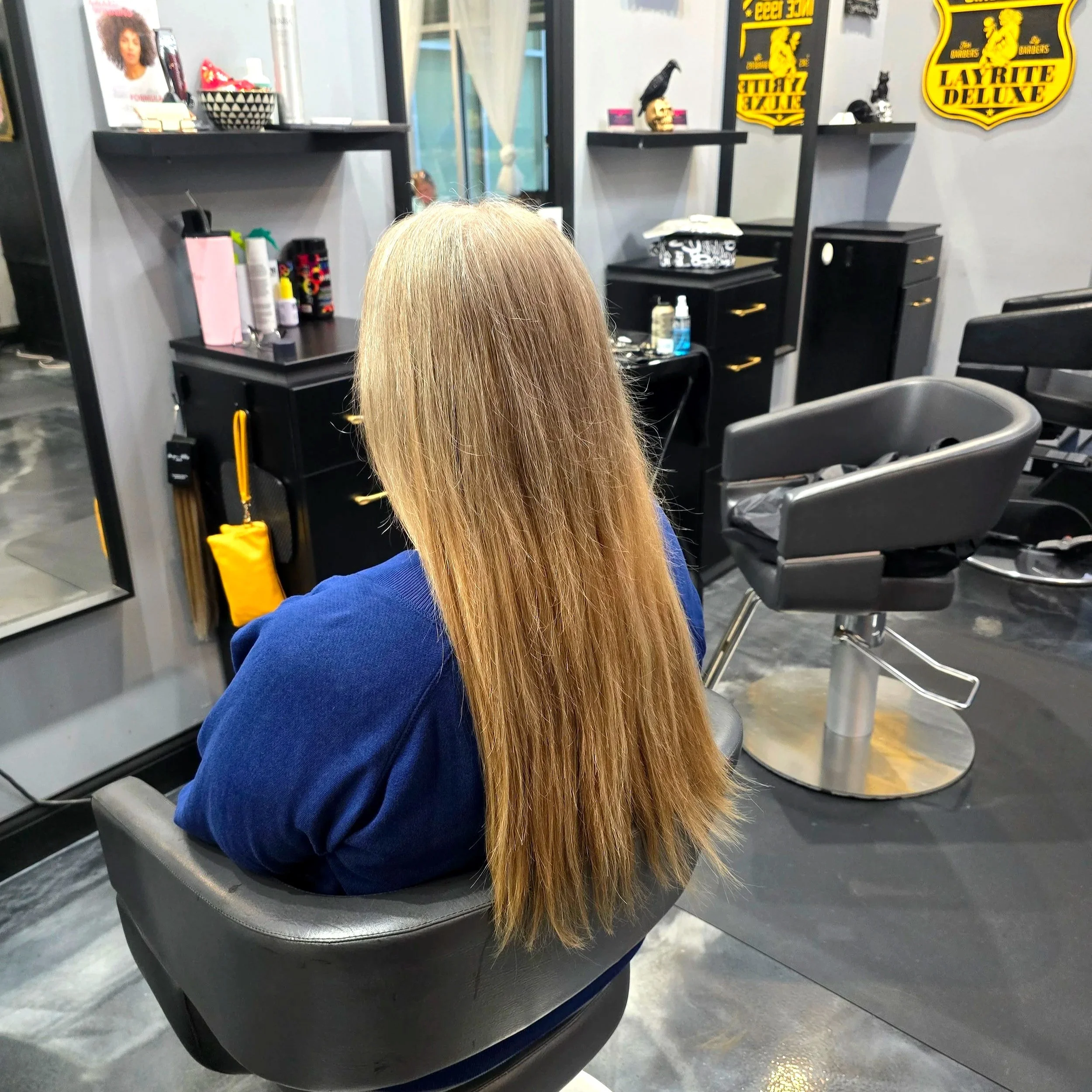 A woman with long blonde hair sitting in a black salon chair inside a hair salon, facing away from the mirror and looking down. The salon has black styling stations with various hair products, a mirror, and décor including black and yellow signs.