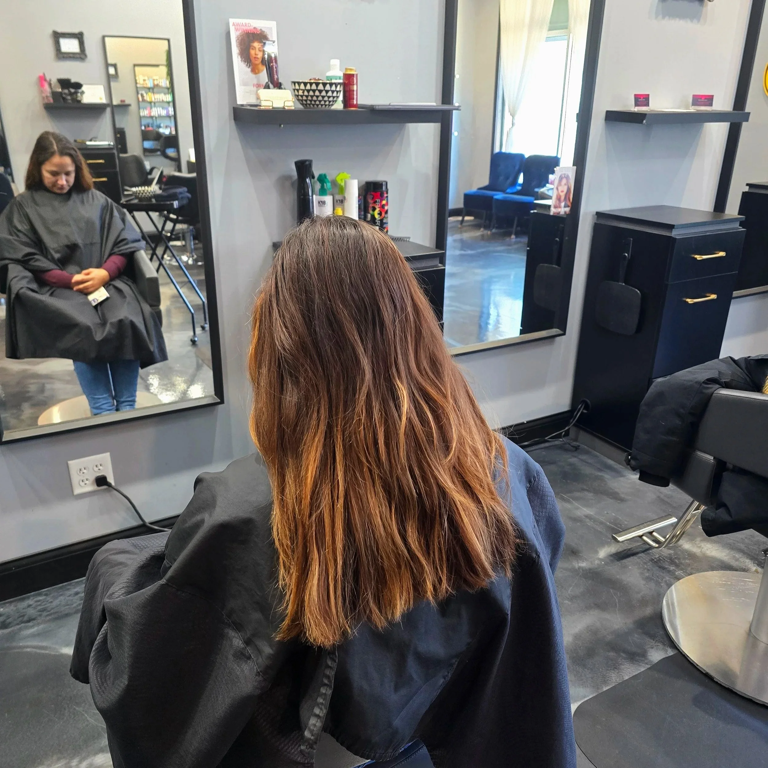 A person with long brown hair and a black salon cape sitting in a hair salon, facing a large mirror. In the reflection, another person with brown hair is sitting in a salon chair, also in a salon cape. The salon has black furniture, mirrors, and shel