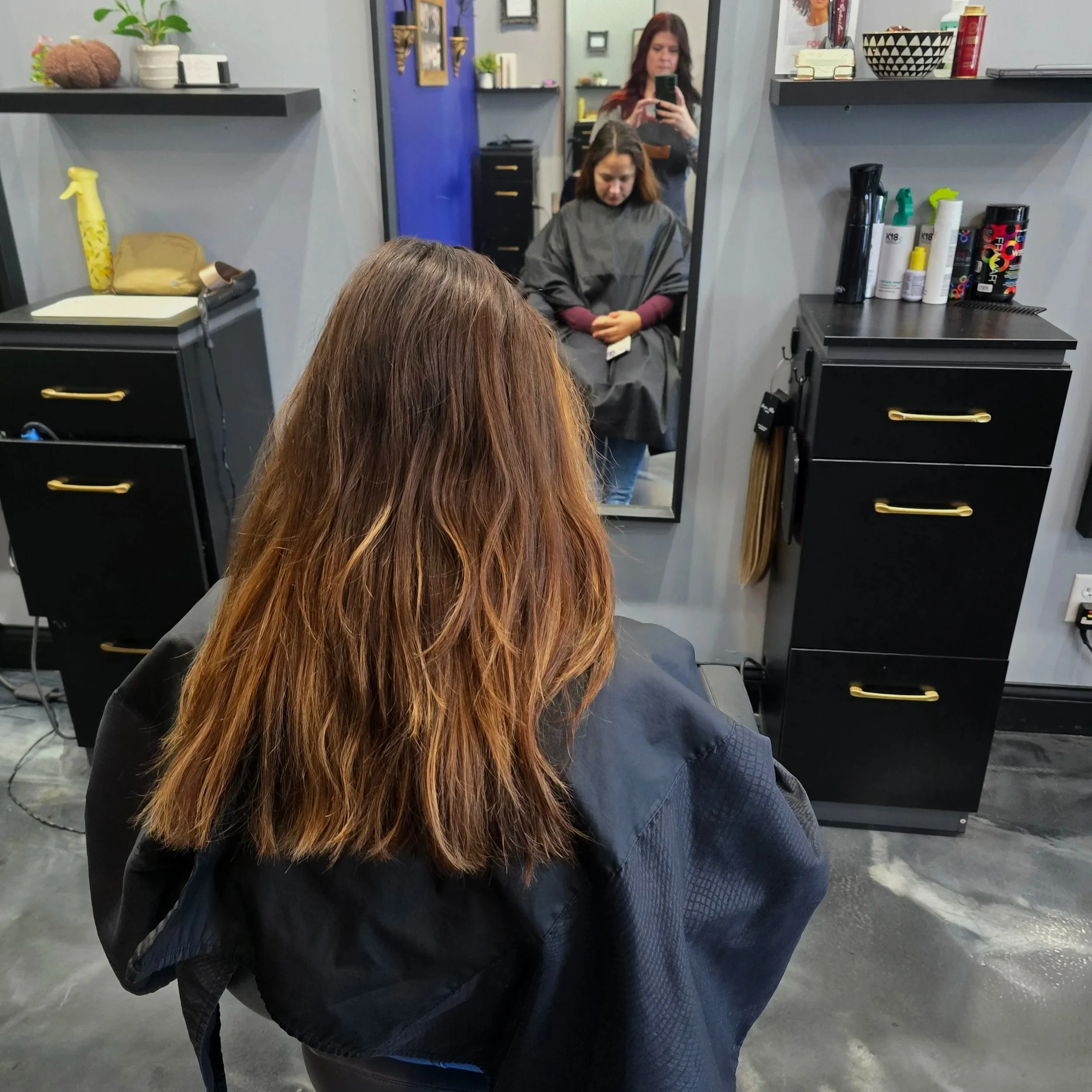 A woman with shoulder-length brown hair sitting in a hair salon chair, facing a mirror. Another woman is taking a photo of her with her phone, and she is wearing a black salon cape. Shelves with hair products and hair tools are visible around the mir