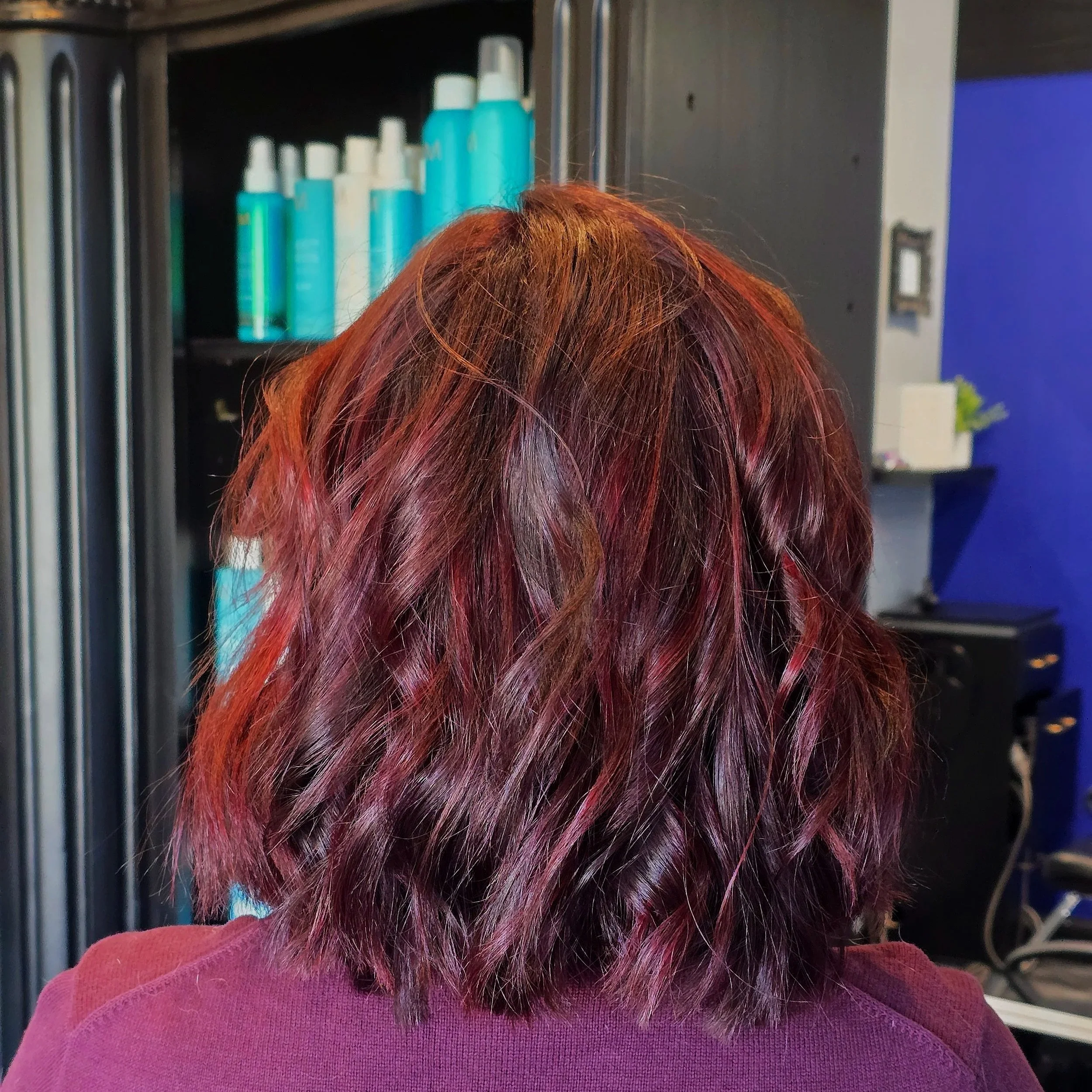 Back view of a woman with shoulder-length, wavy hair dyed in shades of red and purple, inside a salon.