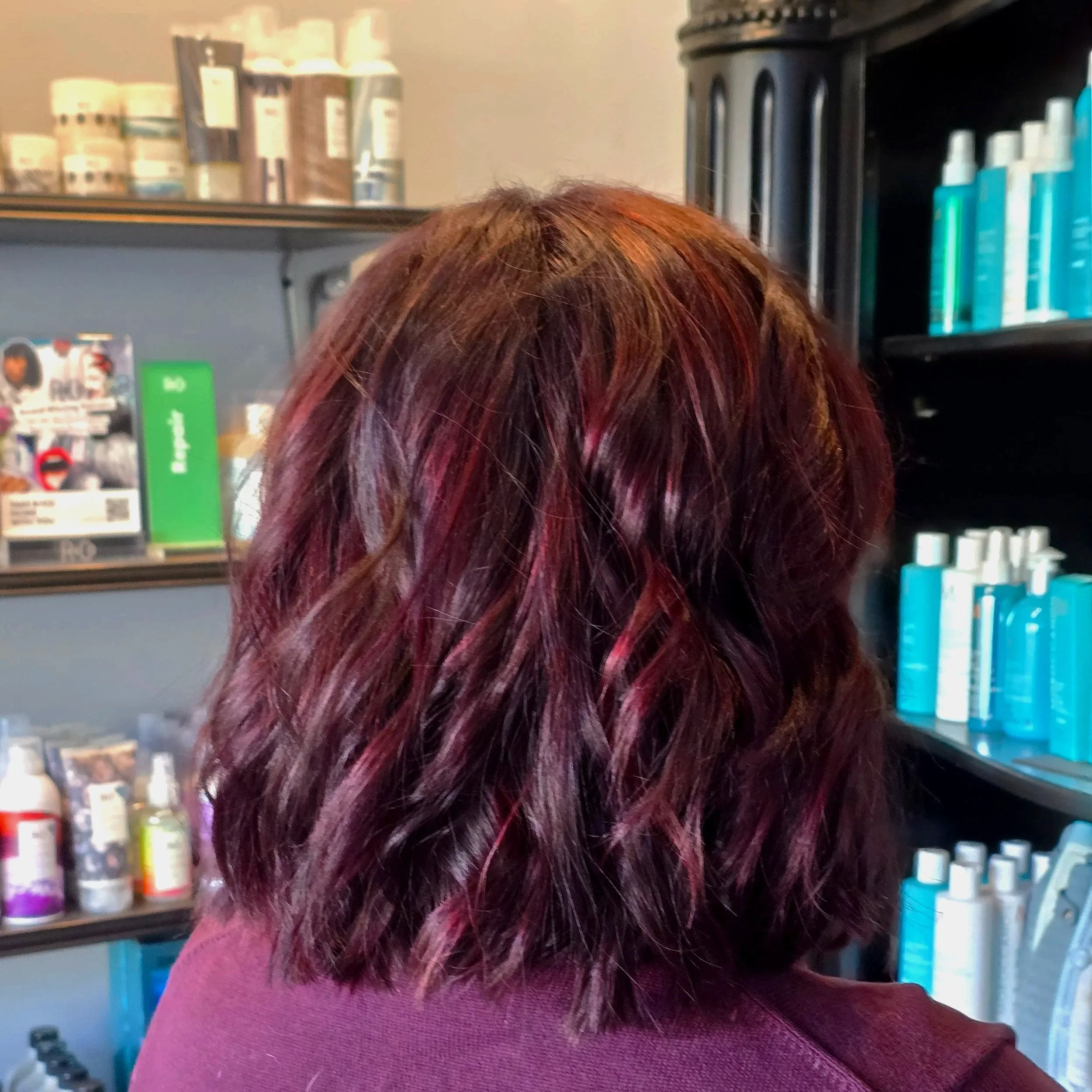 Back view of a person with shoulder-length wavy hair dyed in shades of red, purple, and brown, standing in a salon with shelves of hair and beauty products.