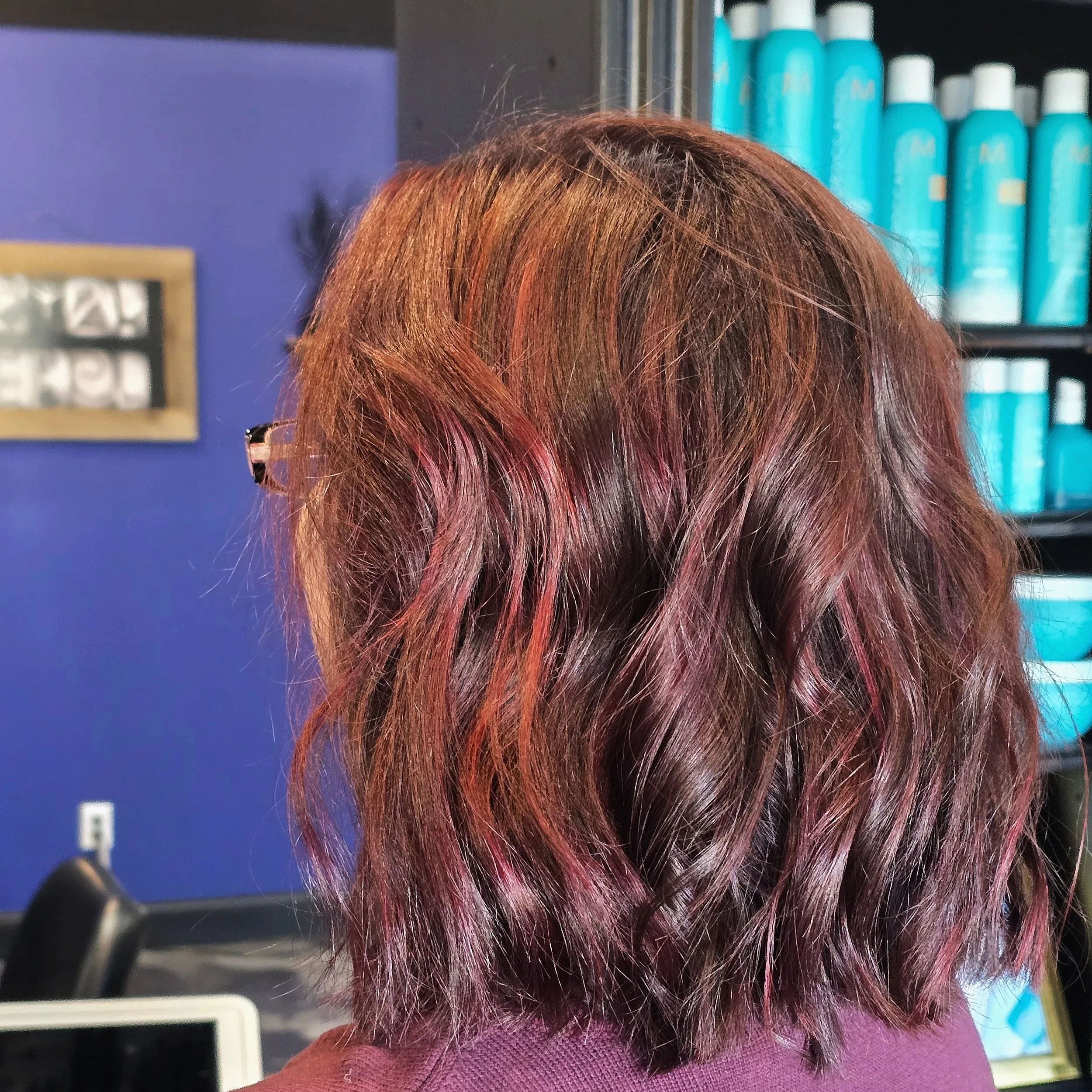 Back of a person's head with wavy, reddish-brown hair with pink highlights, in a salon or hair studio.
