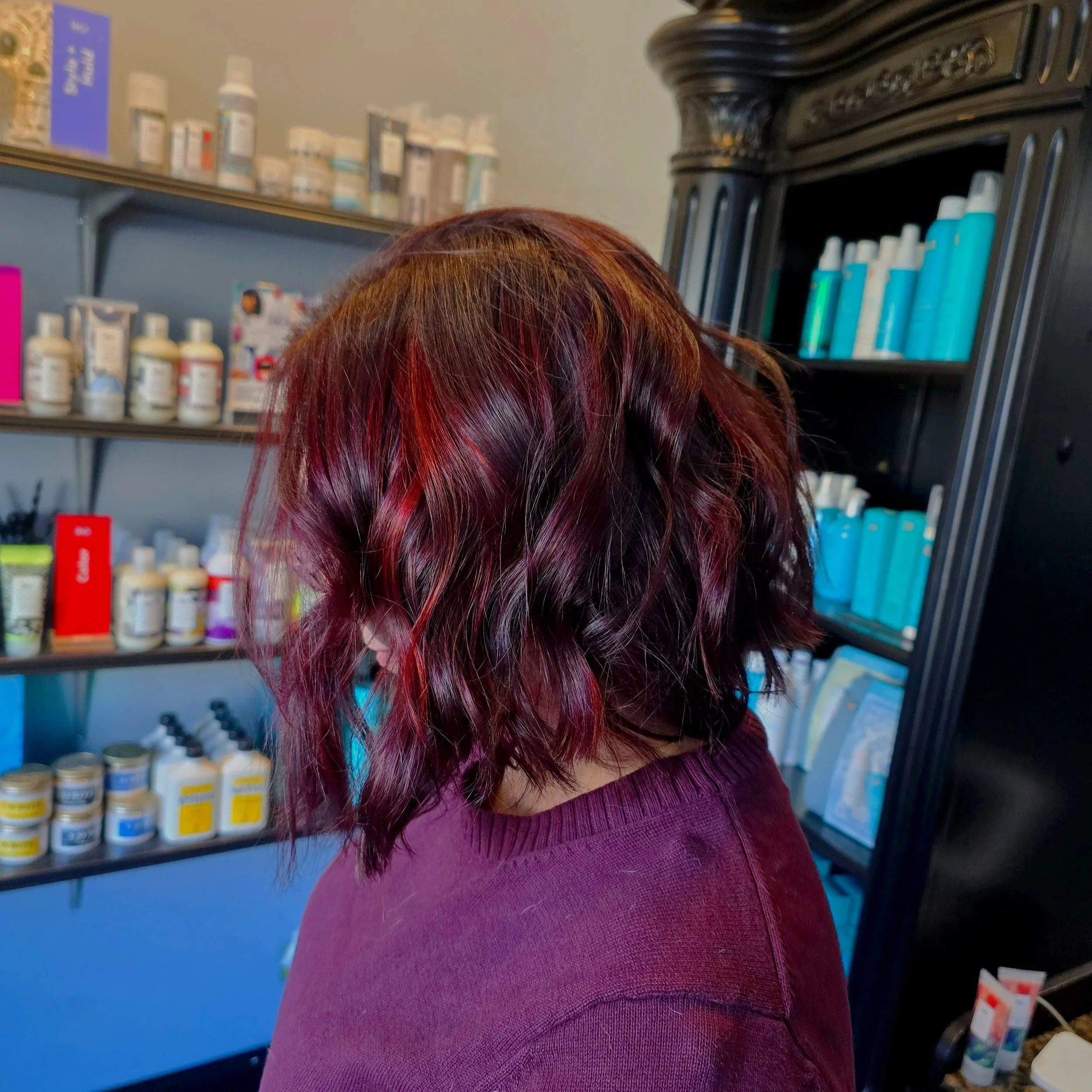 A person with shoulder-length wavy hair dyed in shades of red and purple, wearing a maroon sweater, standing in front of shelves stocked with various bottles and containers, possibly in a pharmacy or beauty supply store.