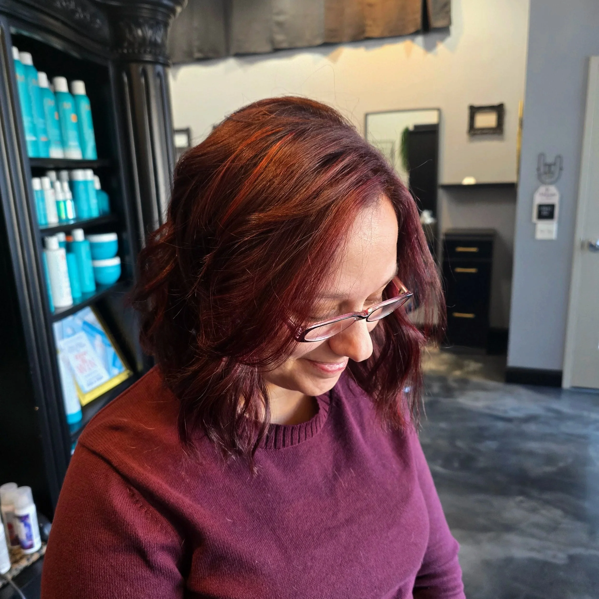 A woman with wavy, burgundy hair and glasses, looking down and smiling, sitting in a salon with shelves of hair products in the background.