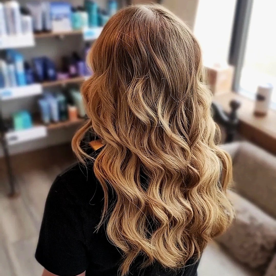 A woman with long, wavy blonde hair styled in loose curls stands inside a salon, with shelves of hair products in the background and a window to the right.