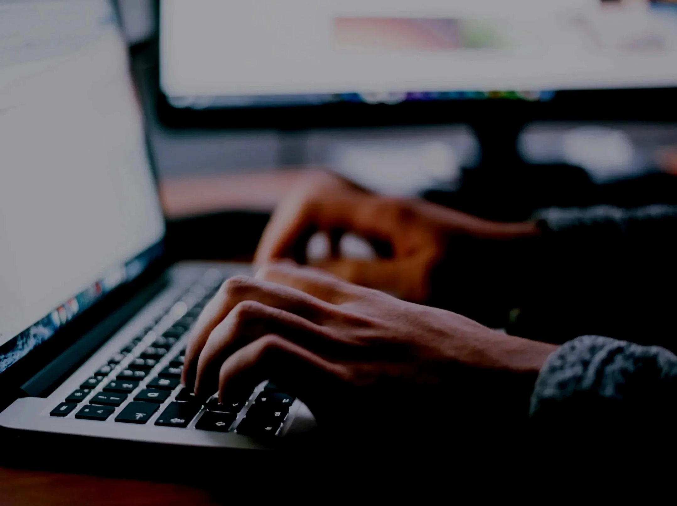 Close-up of hands typing on a laptop keyboard with a blurred computer screen in the background.