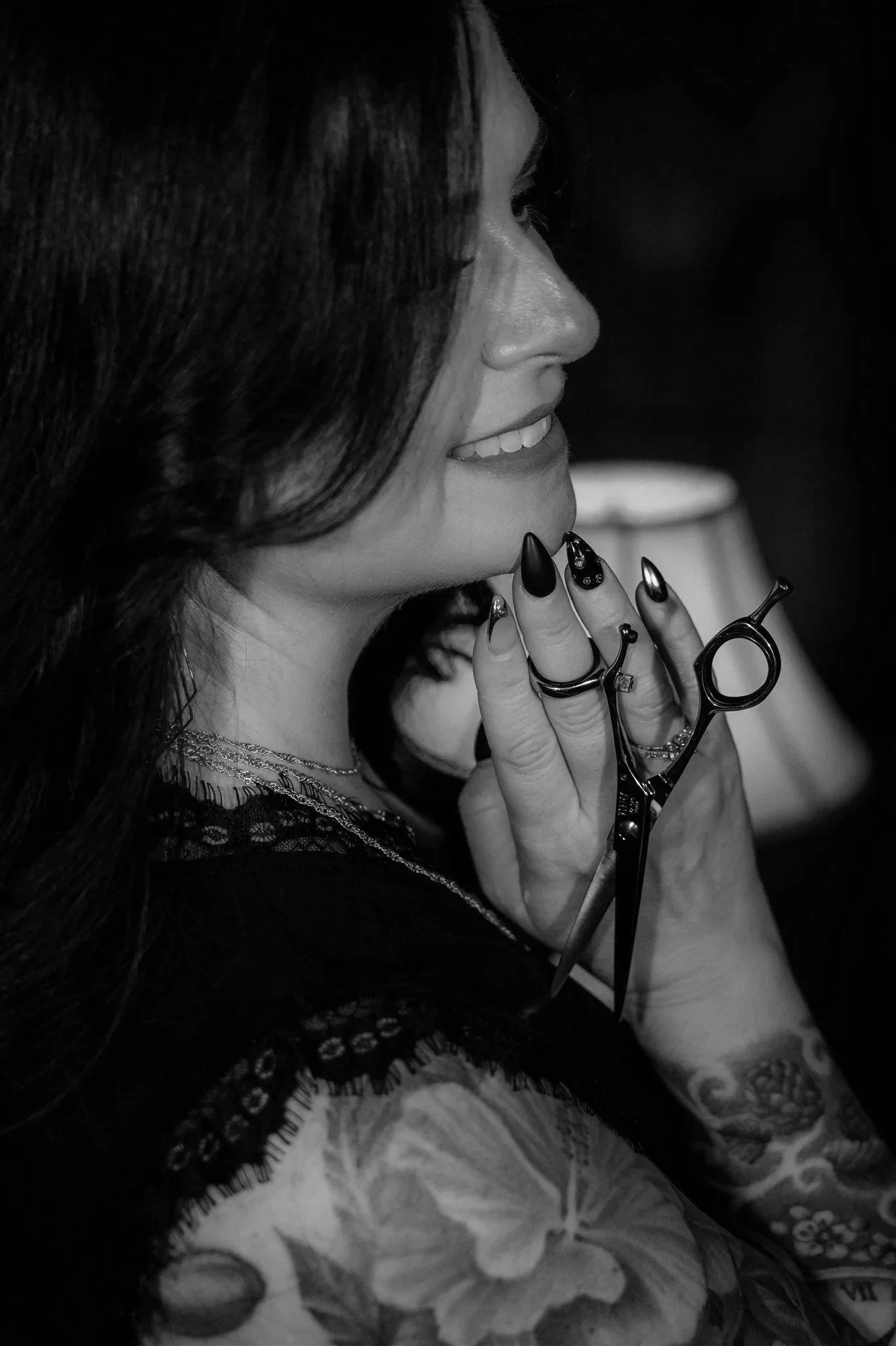 A woman with long dark hair and tattoos on her arm, smiling with closed eyes, holding a pair of scissors near her chin, with a lamp in the background, in black and white.