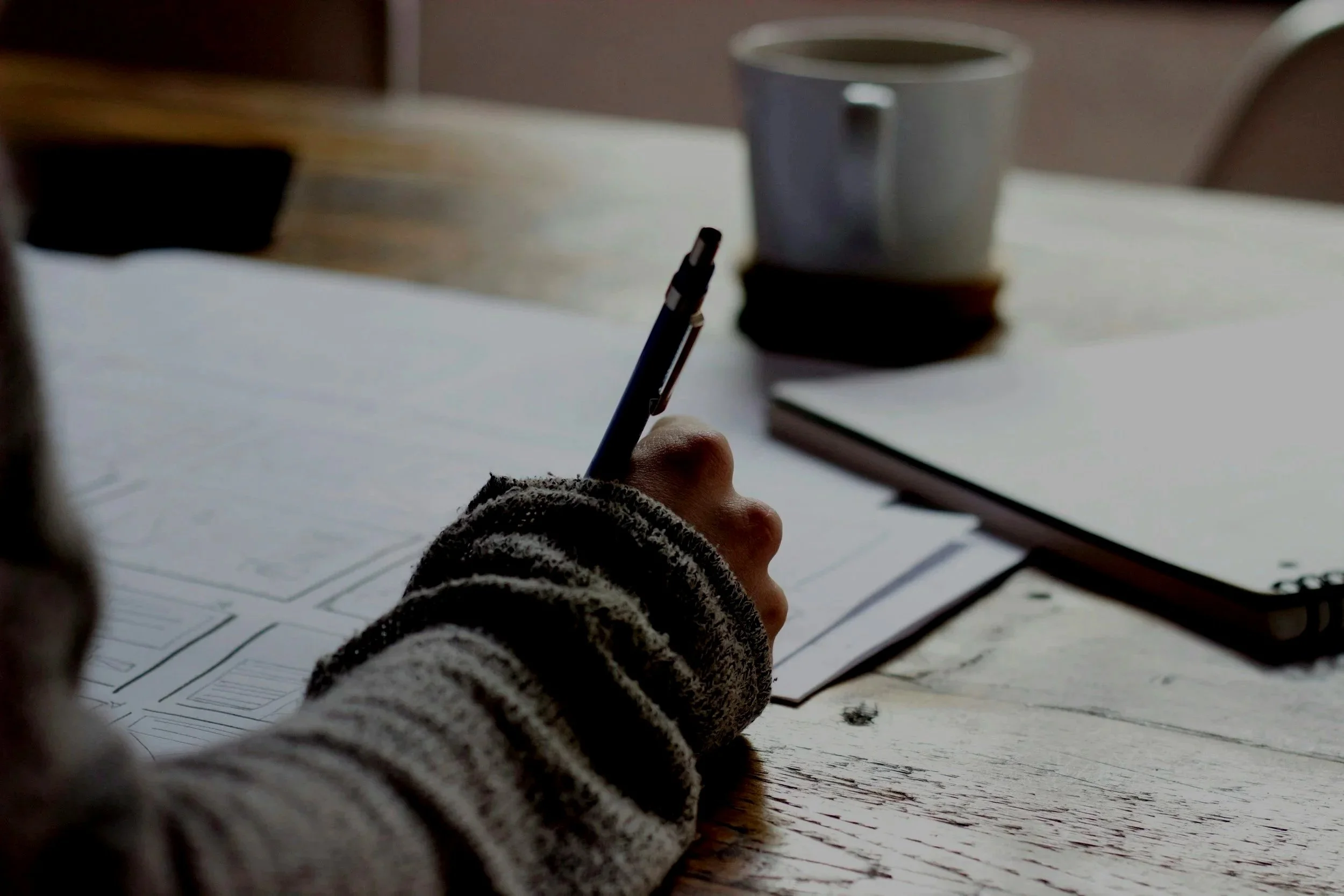 Child writing on paper at a desk with a pen, with notebooks and a coffee cup nearby.