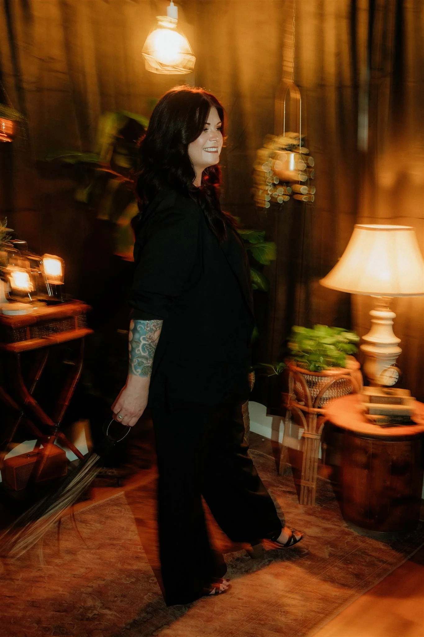 A woman with dark hair and a visible tattoo on her right arm, dressed in black, walking indoors next to a warmly lit lamp and various furniture, with a warm, slightly blurred background.