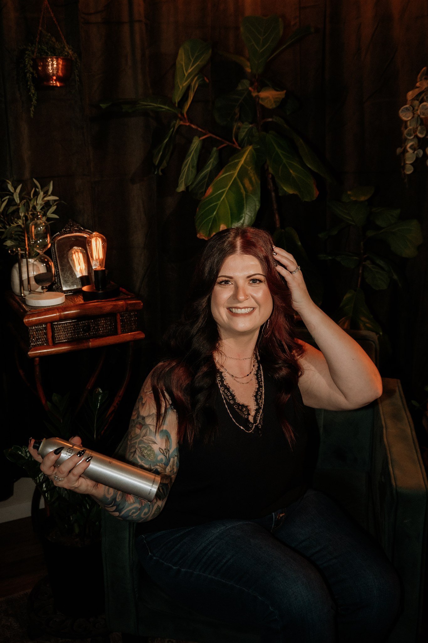 A woman with long dark hair and tattoos on her arm, smiling and sitting on a dark green armchair, holding a can of hair spray in her right hand, in a cozy, dimly lit room with dark wooden walls, large green plants, and warm lighting from vintage-style light bulbs.