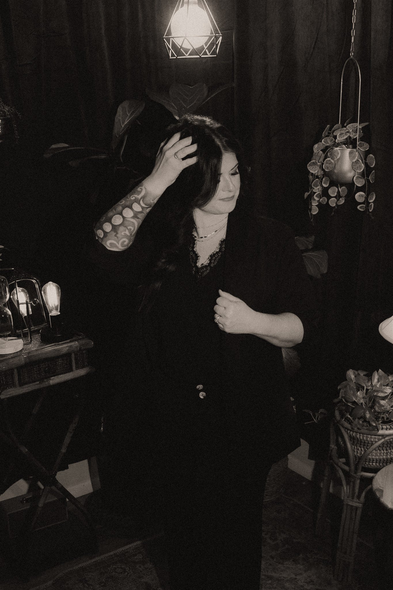 A woman with dark hair and tattoos on her right arm is sitting in a dimly lit room, with her eyes closed and one hand touching her head. There are plants, a hanging light, and various objects around her.
