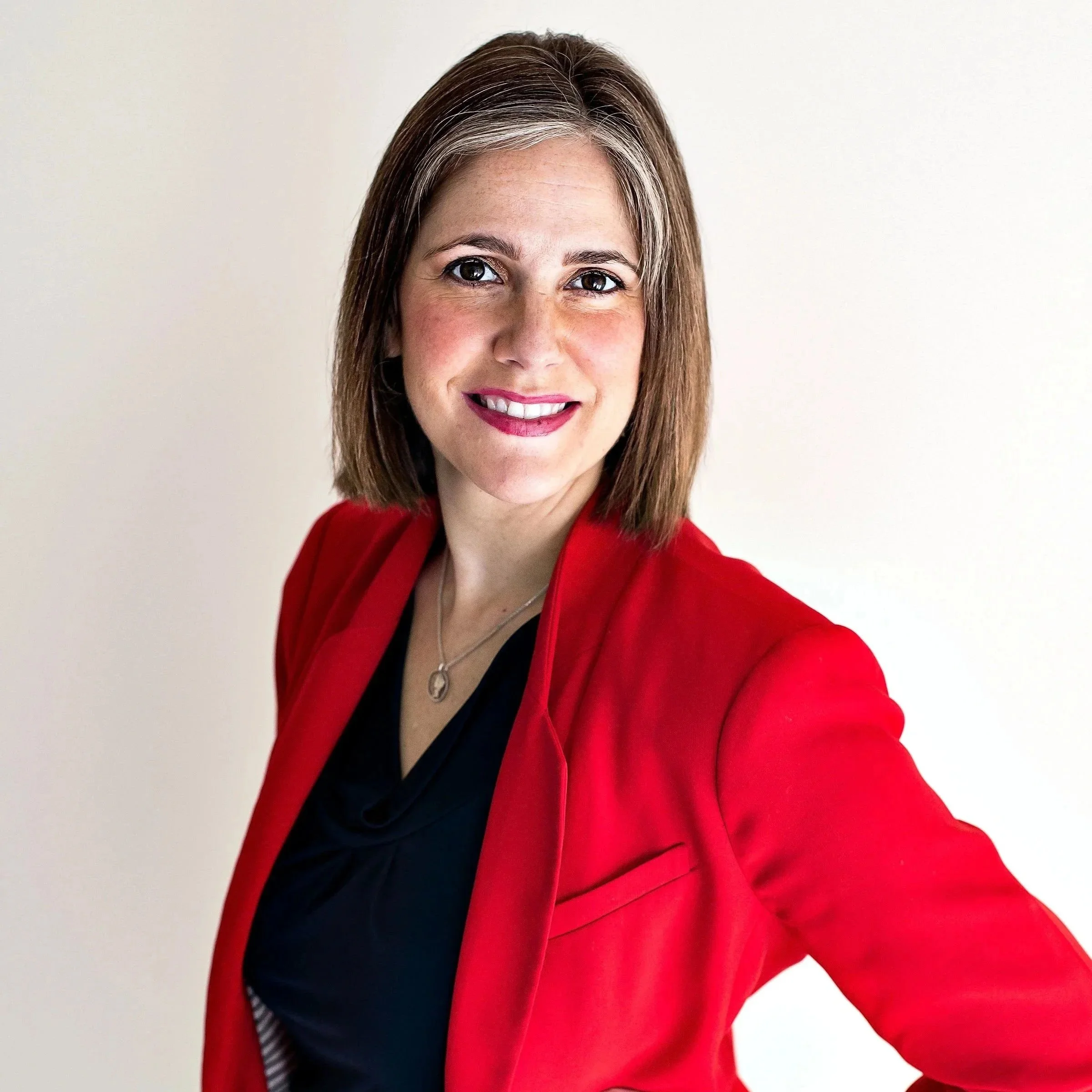 A woman with shoulder-length brown hair, wearing a red blazer and a black top, smiling at the camera against a light-colored background.