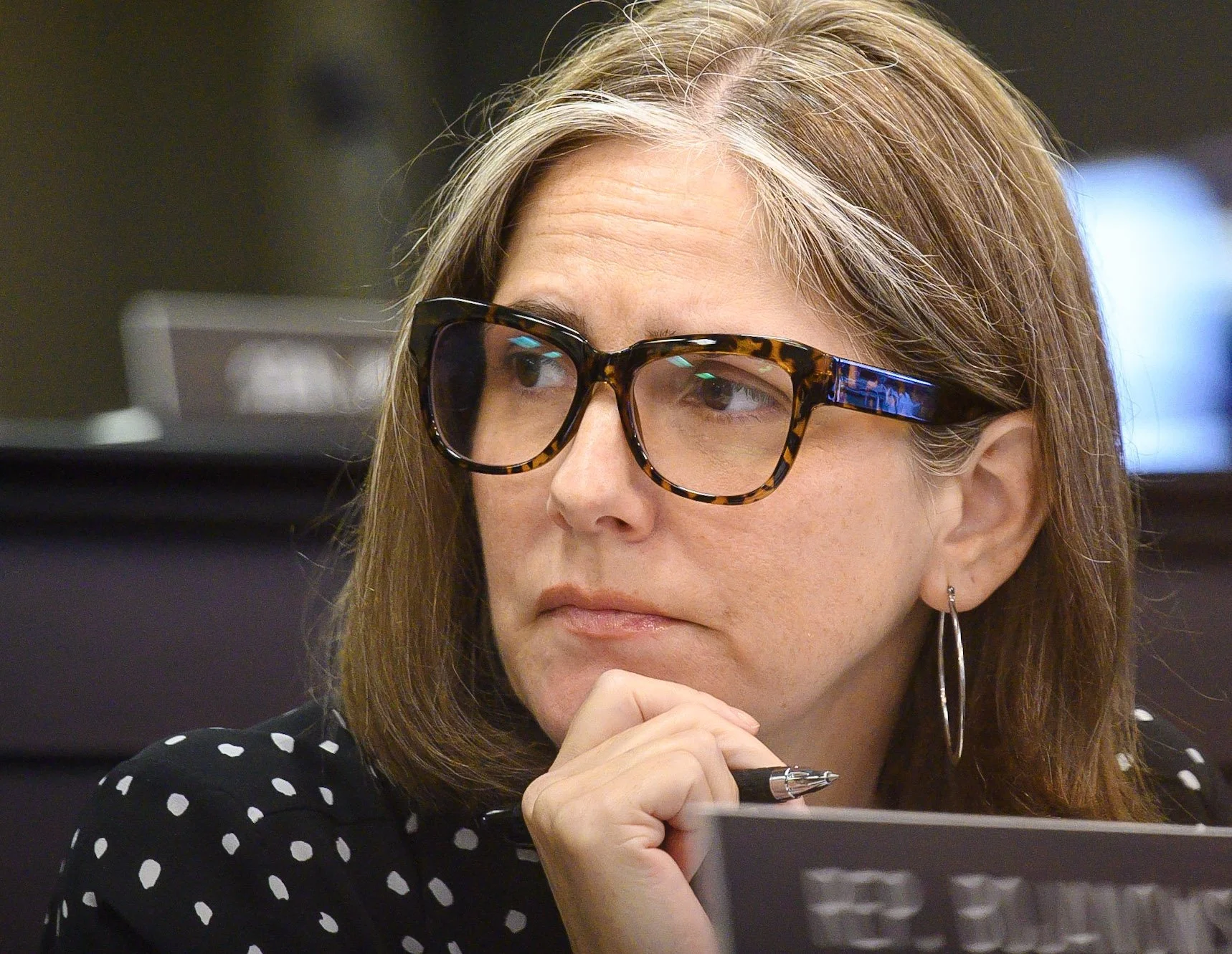 Sarah Stalker, light skin, brown hair, wearing tortoiseshell glasses, silver hoop earrings, a black shirt with white polka dots, and holding a pen, resting her chin on her hand, looking pensively to the side.