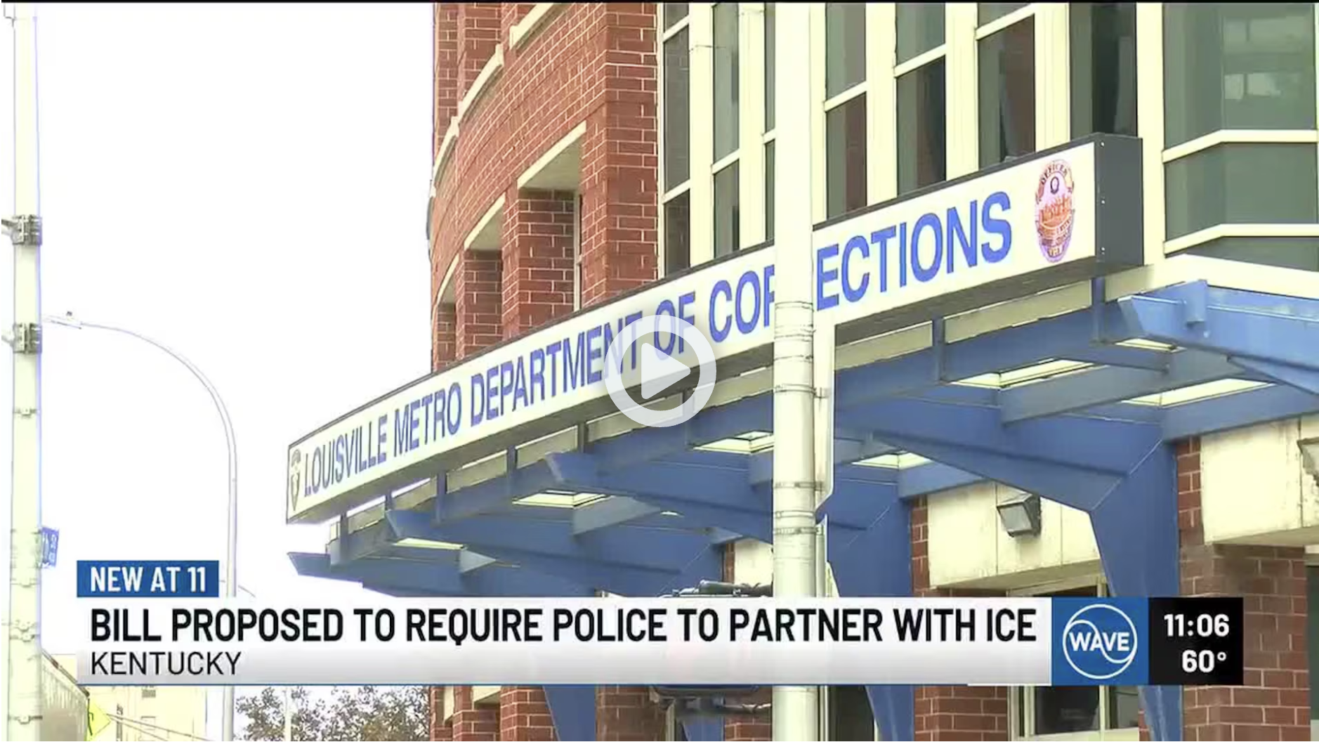 Exterior view of the Louisville Metro Department of Corrections building with a sign above the entrance indicating its name. The building is made of brick with large windows and a blue overhang. News ticker about a bill proposing police partnerships with ICE is displayed at the bottom.