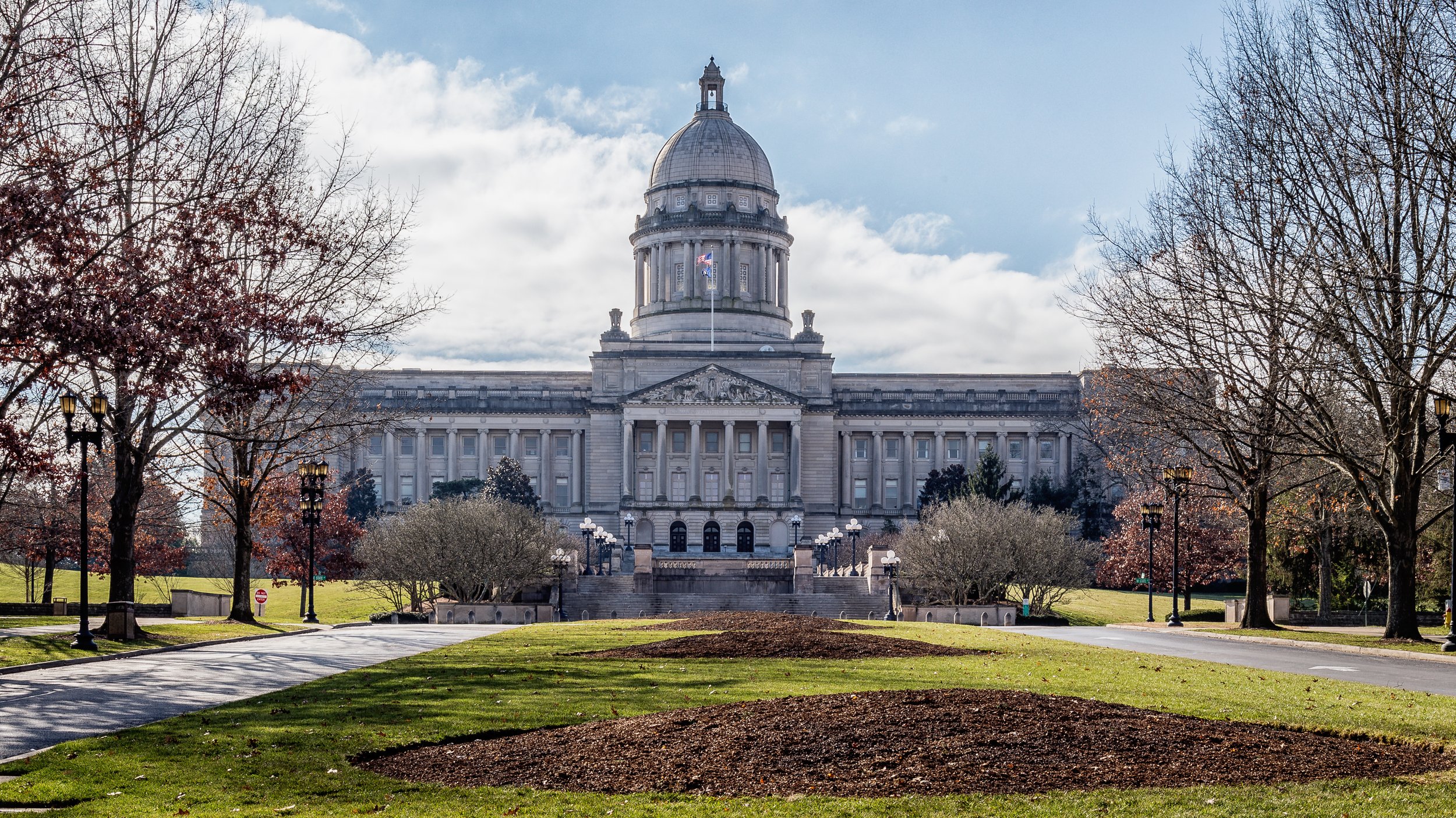 The image features a large, historic government building with a prominent dome, surrounded by bare trees and a well-maintained lawn, under a partly cloudy sky.