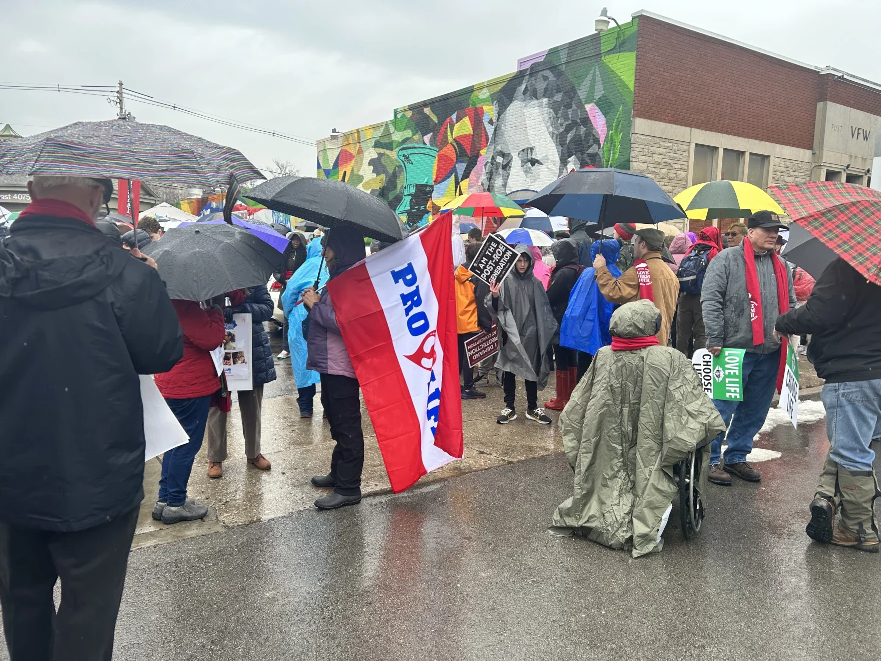 Crowd of people gathered outdoors in rainy weather, many holding umbrellas and protest signs, with a mural mural on a building in the background.