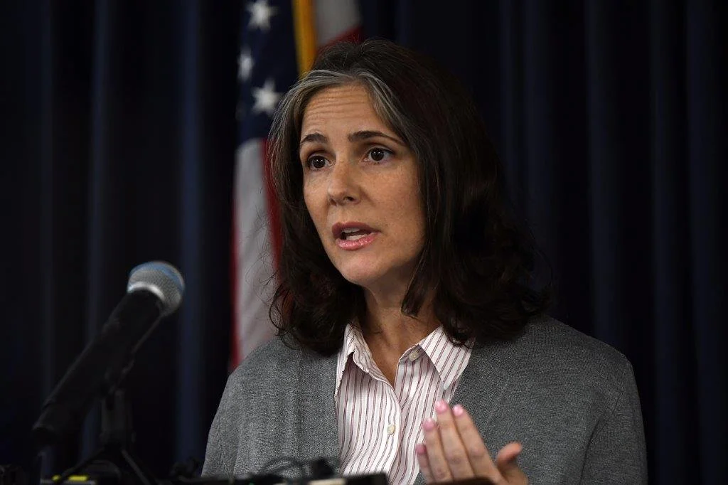 A woman with dark hair speaking at a microphone, gesturing with her hand, standing in front of an American flag and a blue curtain.