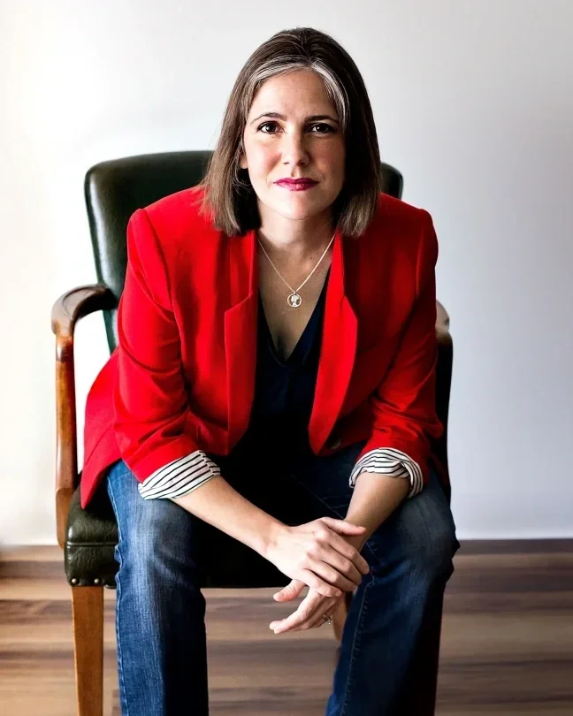 Sarah Stalker, with shoulder-length brown hair, wearing a red blazer, black top, and jeans, sitting on a dark green chair with wooden armrests in a room with a white wall and wooden floor.