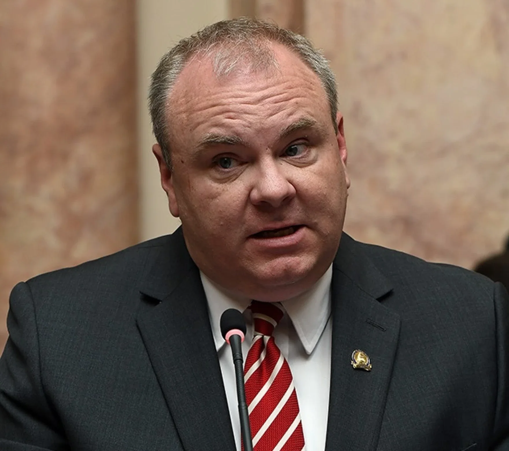 A middle-aged man with short, receding gray hair, wearing a dark suit, white shirt, red and white striped tie, and a lapel pin, speaking into a microphone against a brown marble background.