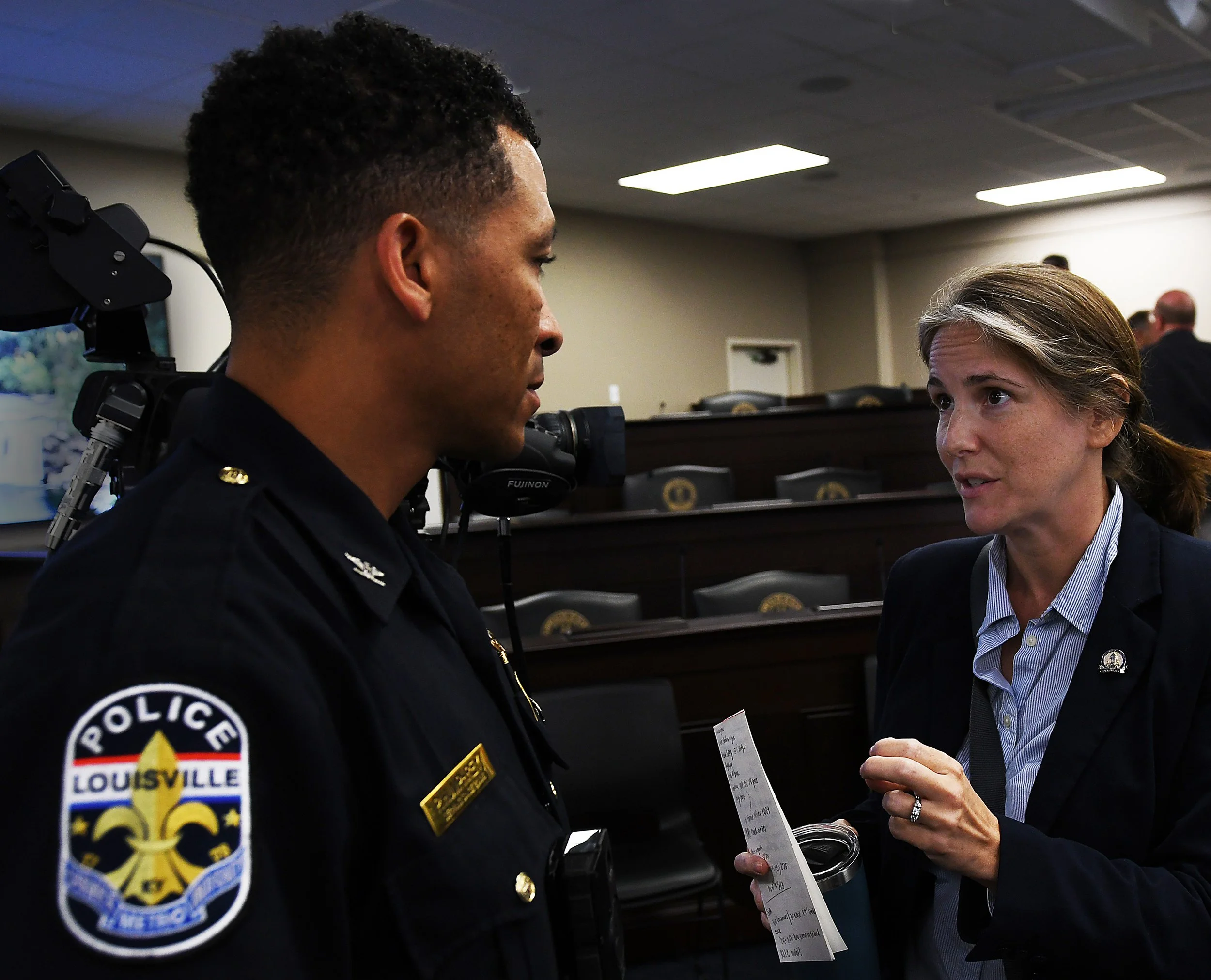 A police officer and Sarah Stalker engage in a conversation, with the woman holding a piece of paper and a water bottle, in a conference room or courtroom setting.