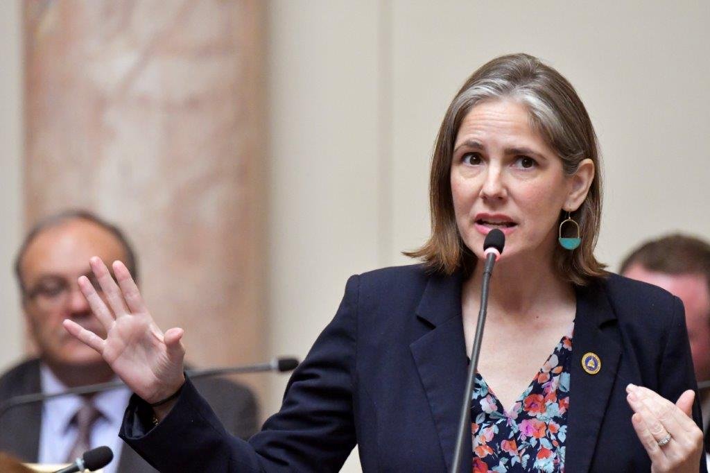 Sarah with shoulder-length brown hair, wearing a dark blazer, floral blouse, and turquoise earrings, is speaking into a microphone during a formal event. She is gesturing with her left hand, and there are people sitting behind her in the background.