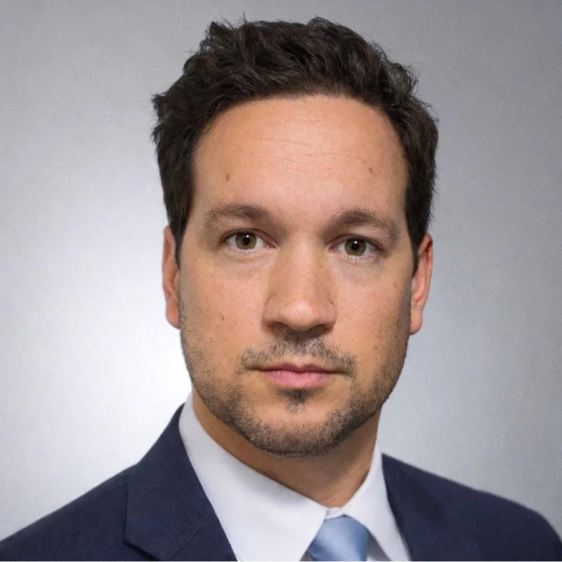 Close-up portrait of a man with short dark curly hair, light skin, wearing a dark suit, white shirt, and light blue tie against a plain gray background.