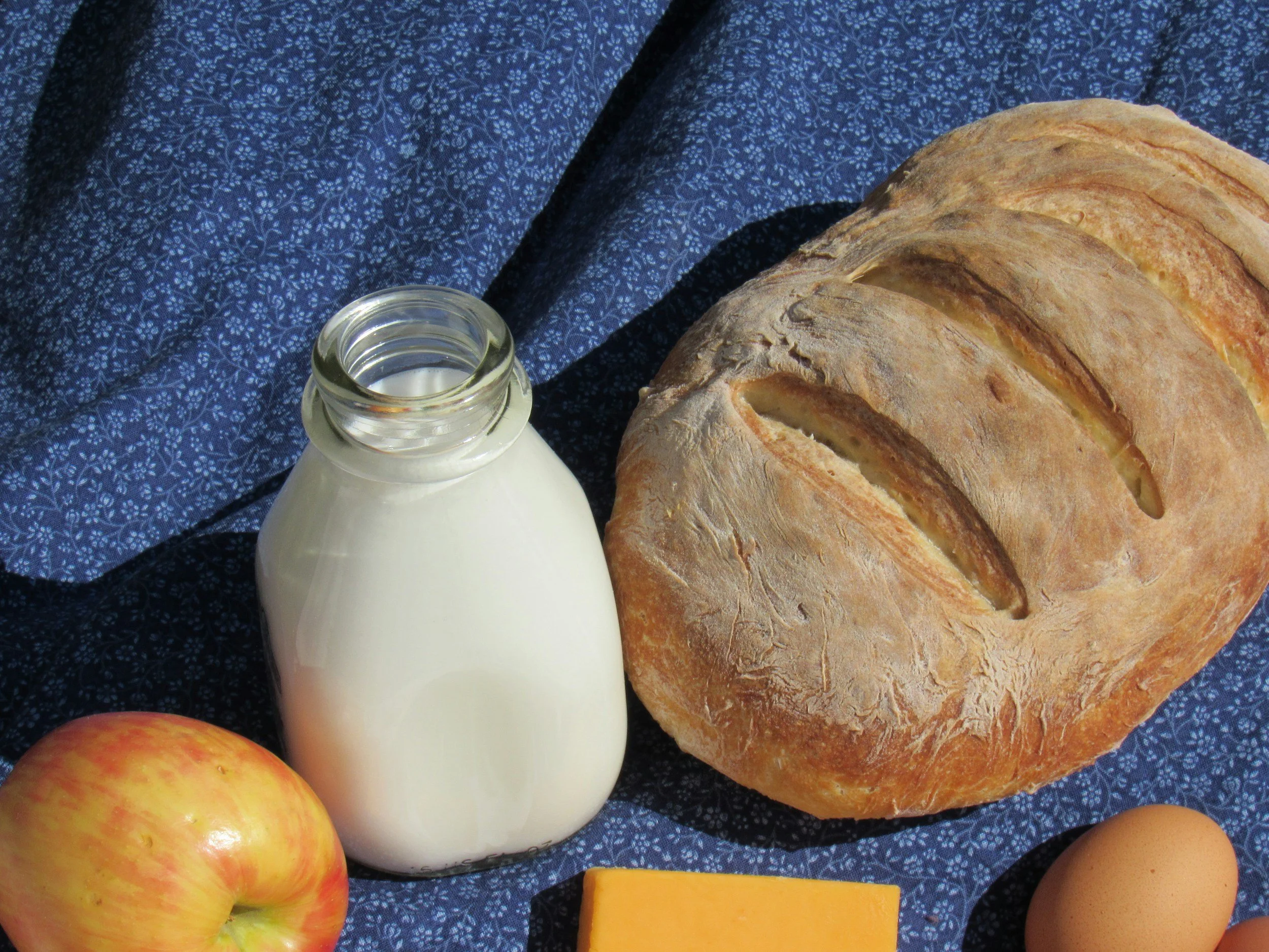 A loaf of bread, a bottle of milk, an apple, a slice of cheese, an egg, and a small yellow fruit or vegetable on a blue patterned cloth.