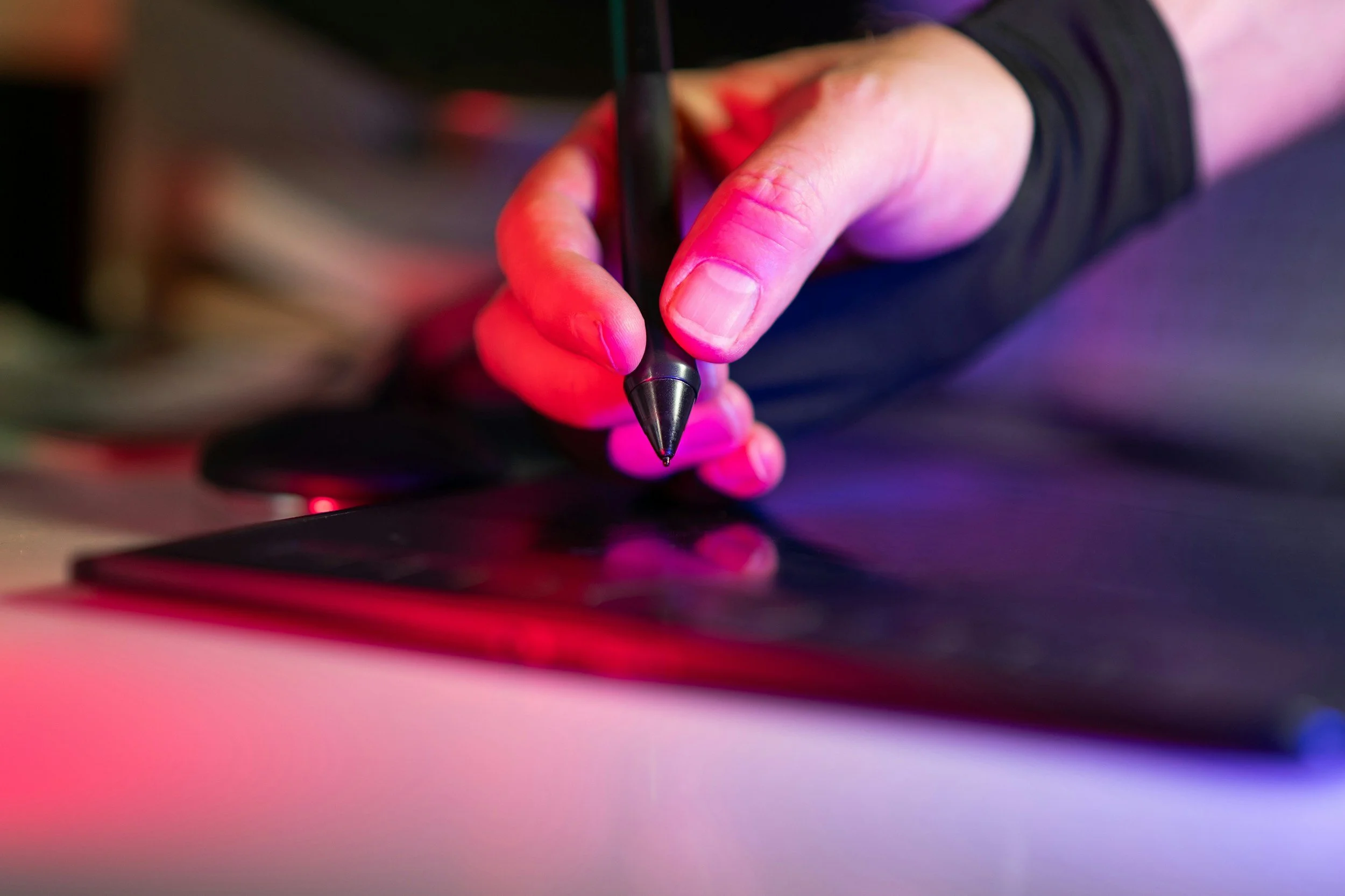 A hand holding a stylus pen over a graphic tablet, with colorful pink, purple, and blue lighting