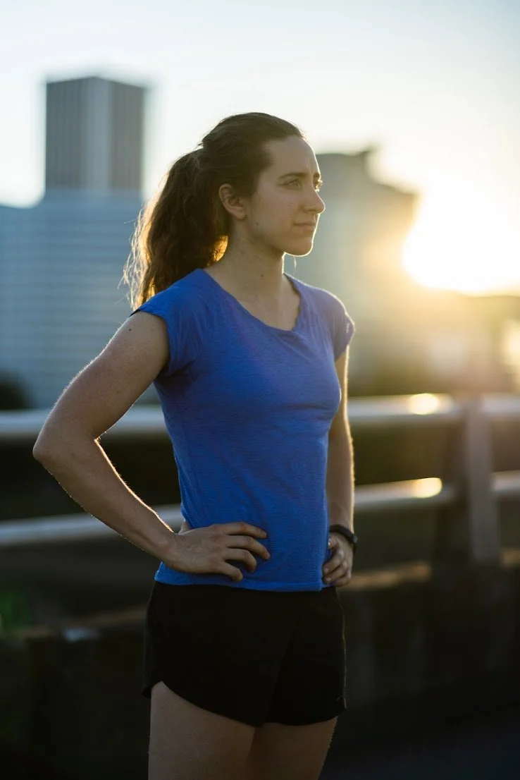 A woman standing outdoors with hands on her hips, wearing a blue athletic shirt and black shorts during sunset in an urban setting.