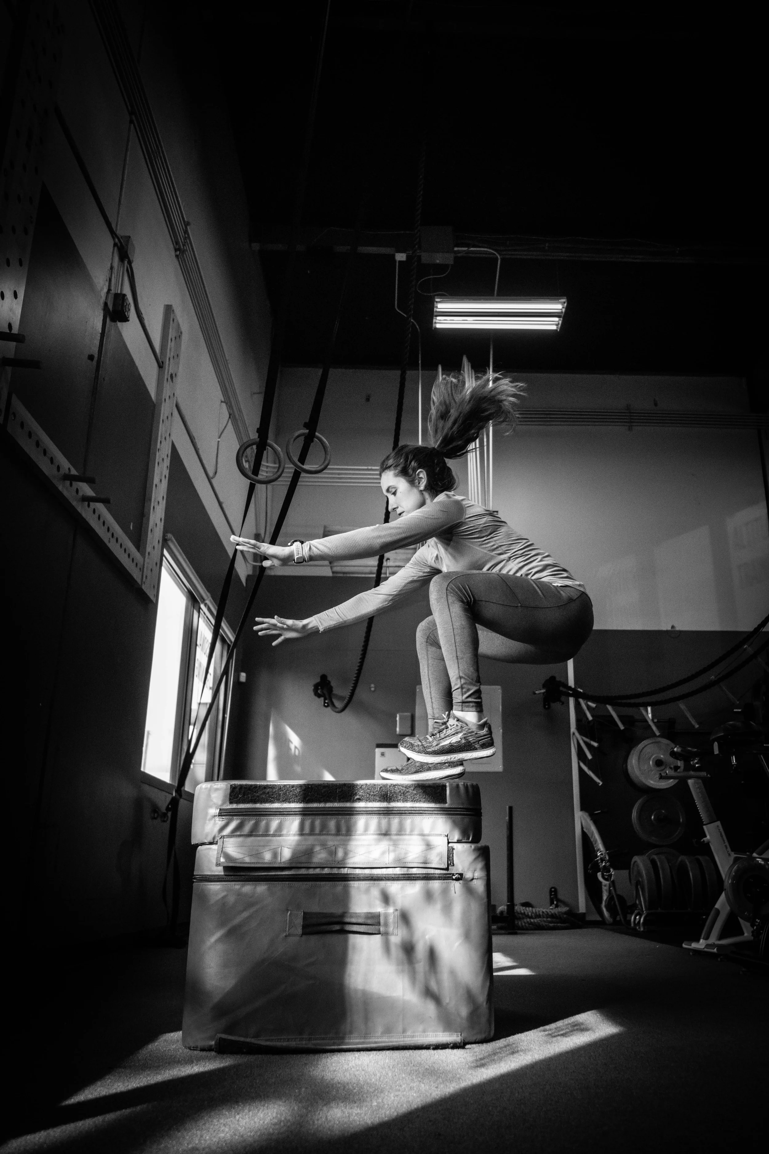 A woman in athletic clothing jumping onto a padded box in a gym, captured in black and white.