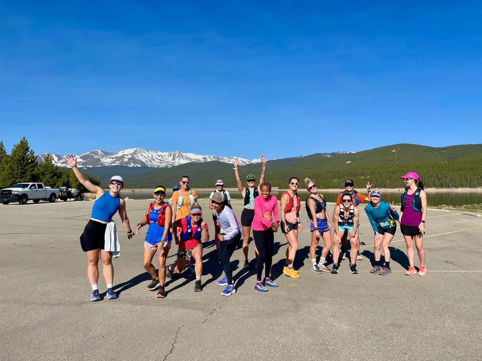 Group of people in outdoor athletic clothing posing on a mountain backdrop with snow-capped mountains, green forests, and a lake.