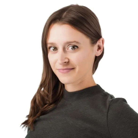 A woman with long brown hair, wearing a dark gray top, smiling slightly, against a plain white background.