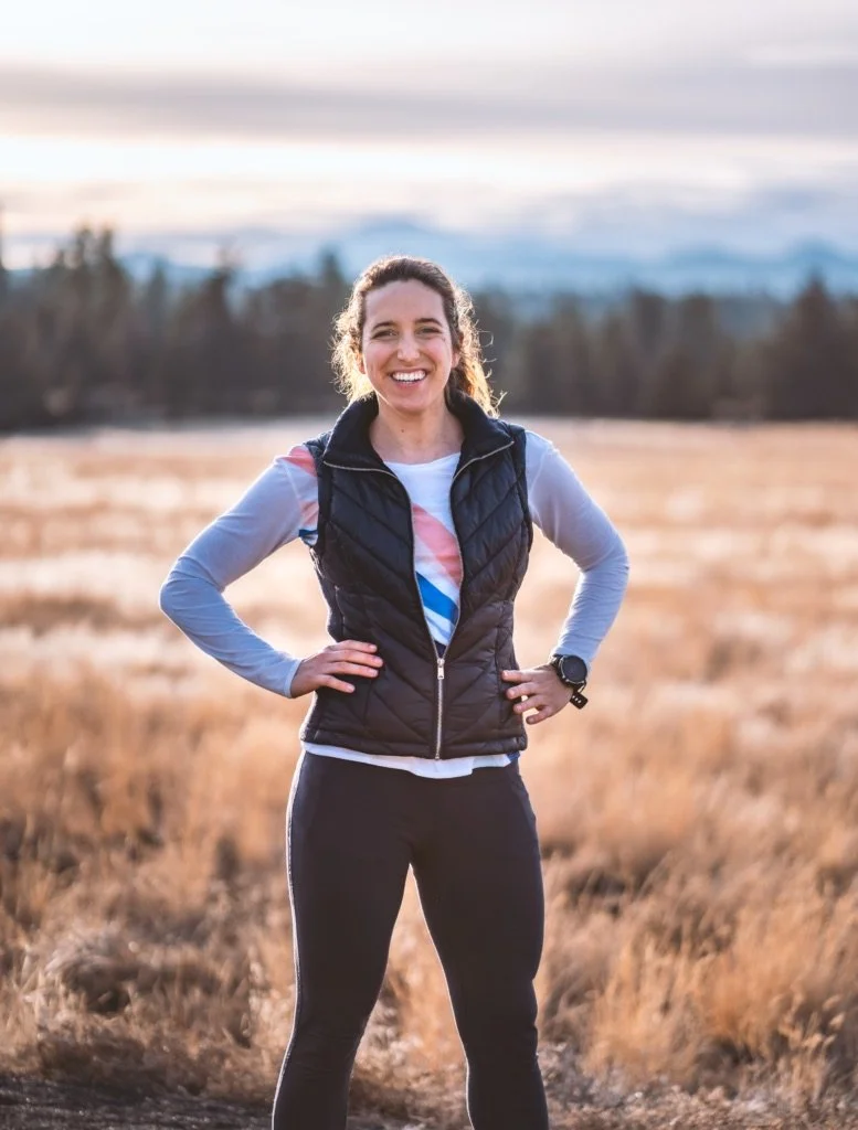 Woman standing outdoors in a field, smiling with hands on hips, wearing athletic clothing and a black vest.