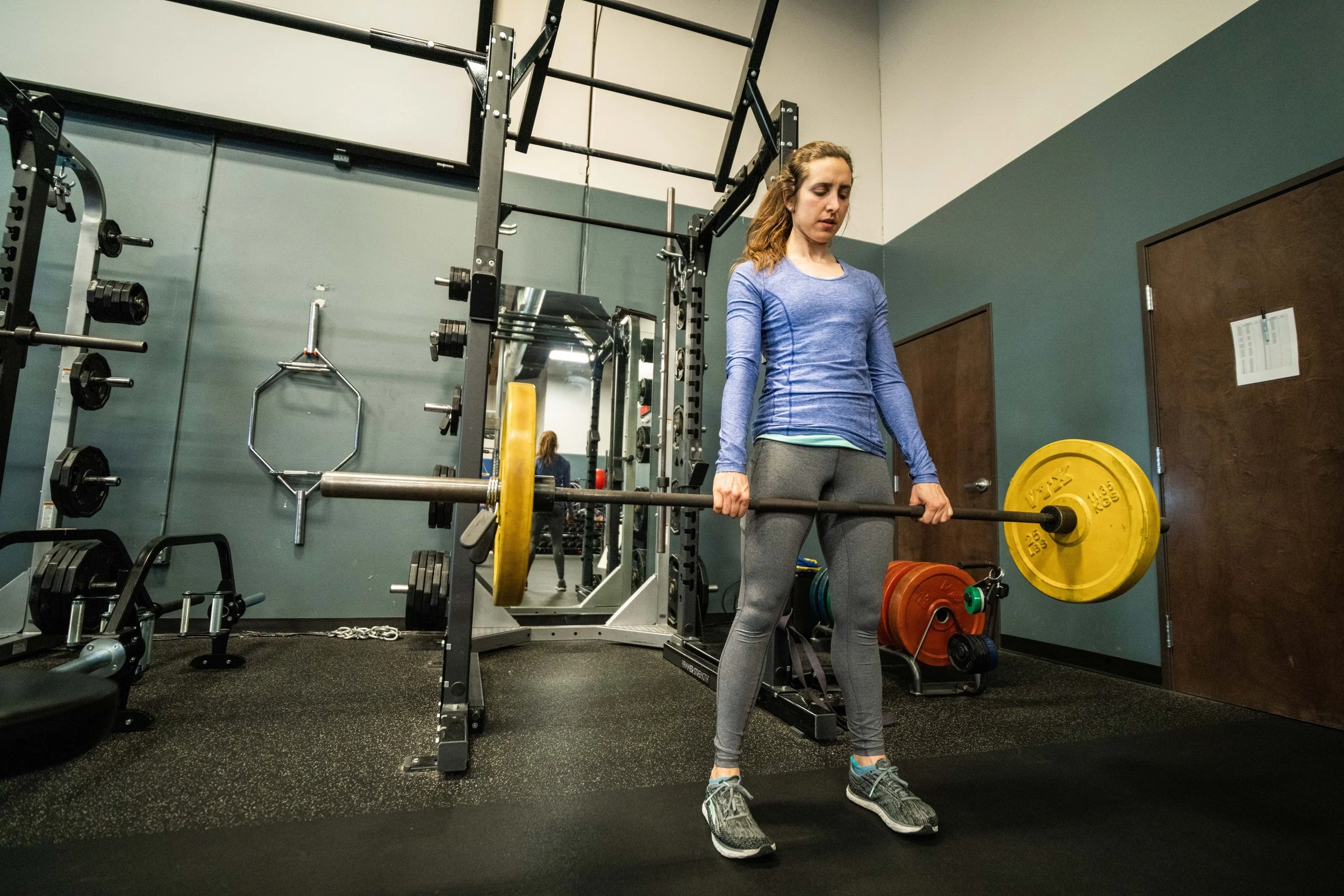 A woman lifting a barbell with yellow weight plates in a gym. She is wearing a blue long-sleeve shirt, gray leggings, and running shoes, standing on a black rubber gym floor.