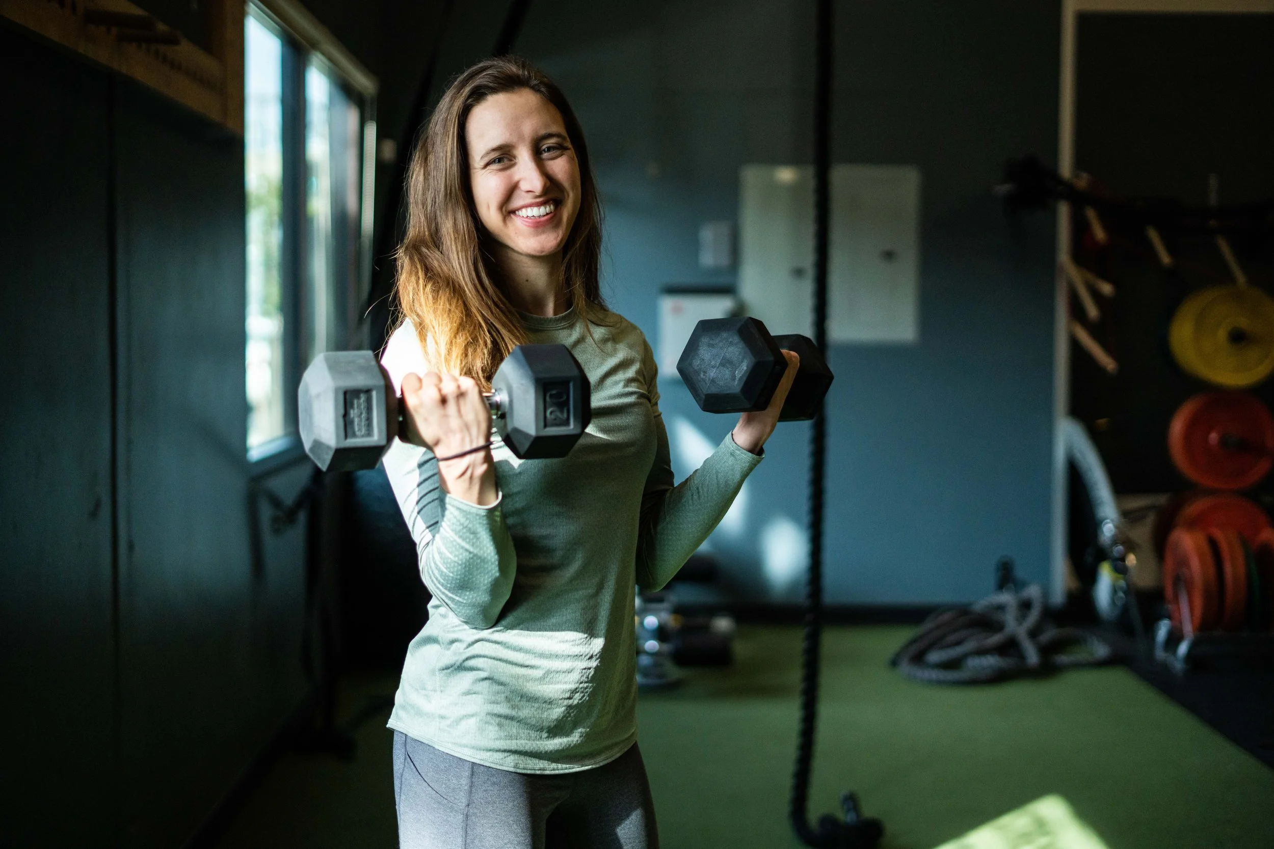 A young woman smiling and holding two dumbbells in a gym.