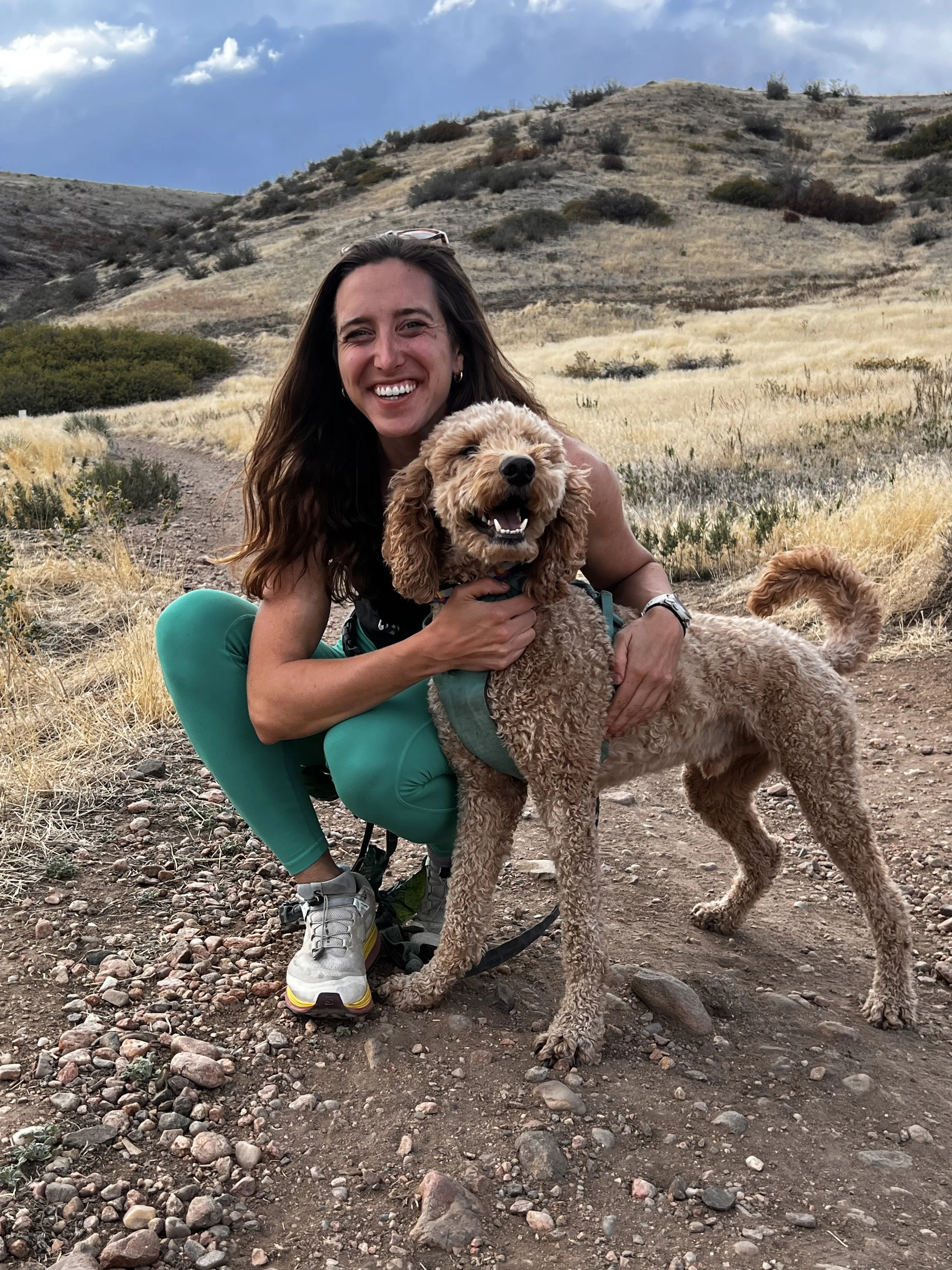 A woman smiling and squatting on a dirt trail with her curly-haired dog, both outdoors in a dry, mountainous landscape with sparse vegetation.