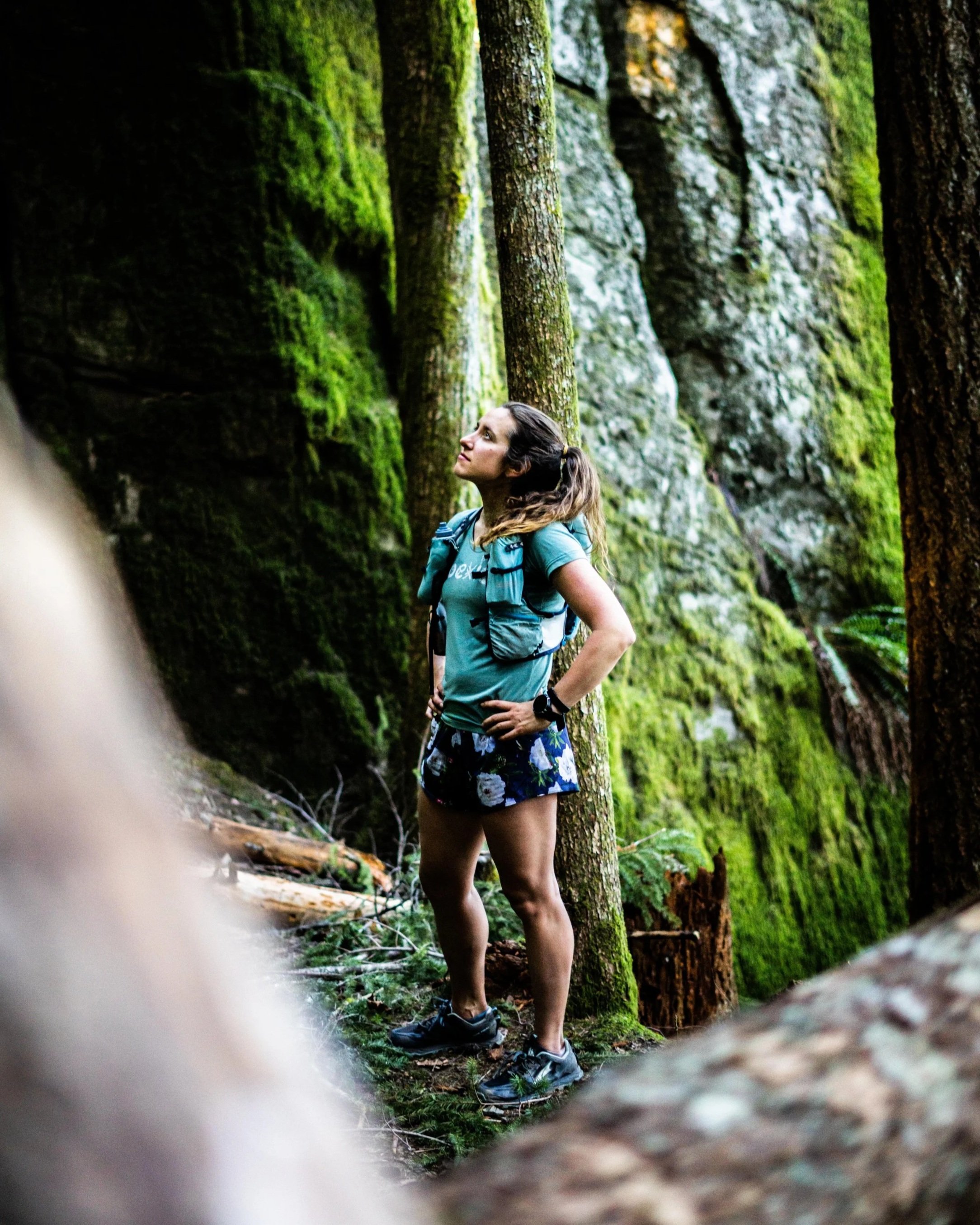 A woman stands in a lush, mossy forest, looking up with an expression of awe. She is dressed in outdoor gear, including a teal shirt, floral shorts, and hiking shoes, with a backpack and smartwatch.