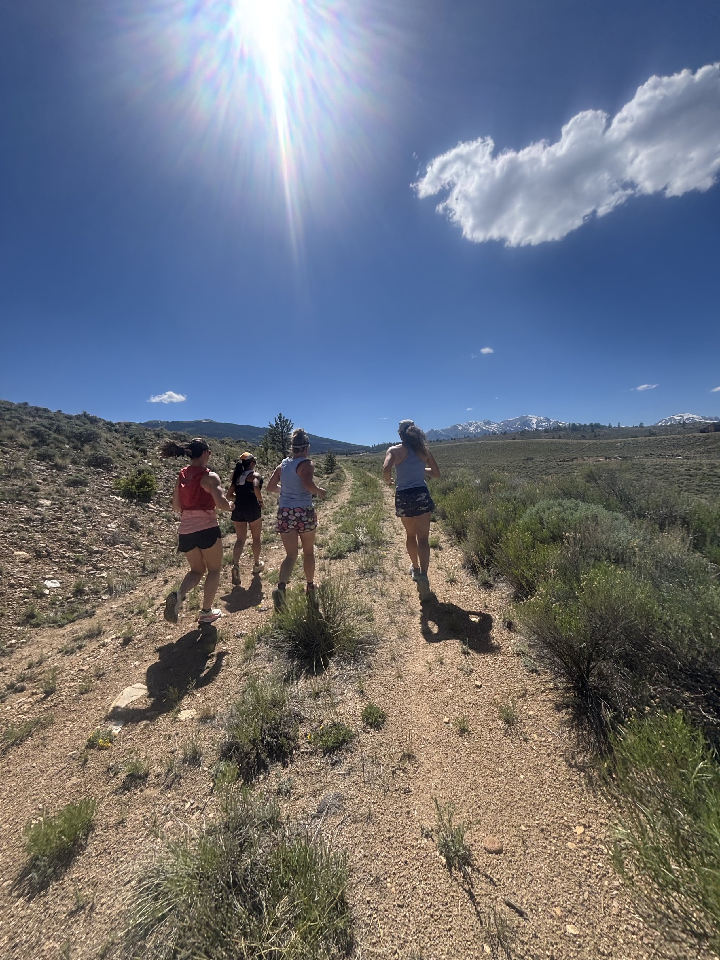 Five women jogging on a dirt trail in a mountainous area with sparse vegetation under a bright sunny sky with a few clouds.
