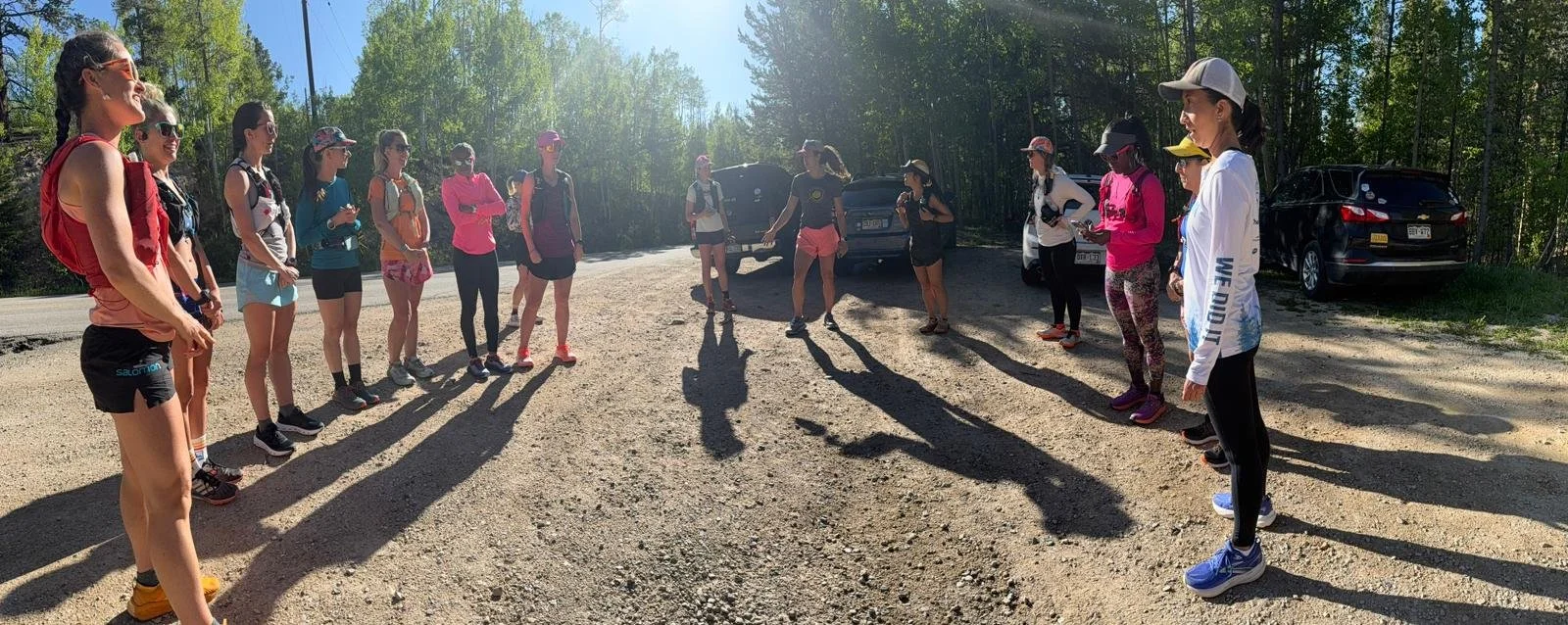 A group of women gathered outdoors on a dirt road in a wooded area, standing in two lines facing each other with a woman speaking in the middle. They are dressed in athletic clothing and appear to be preparing for a running or hiking activity.