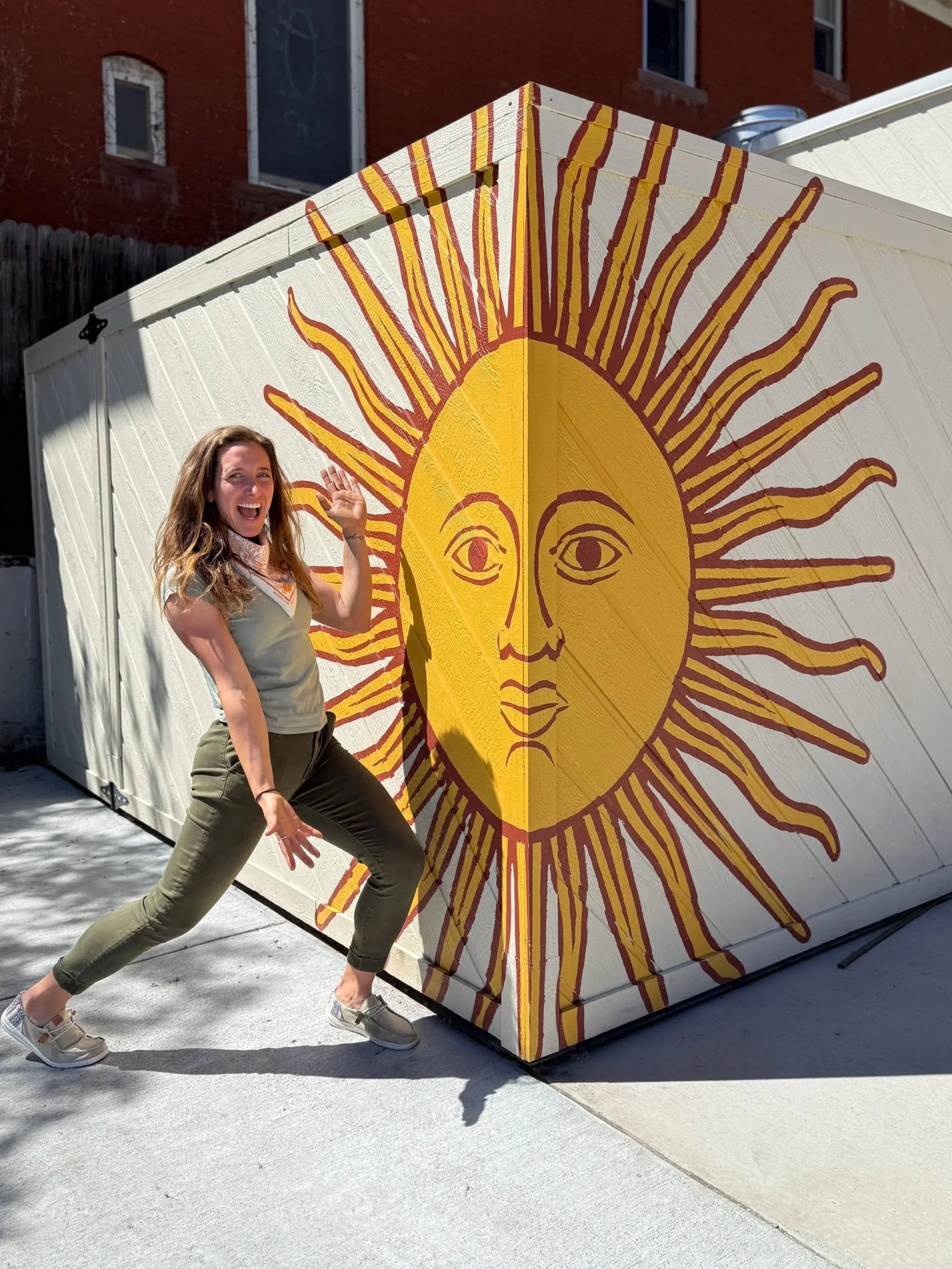 A woman standing in front of a white shed with a large sun face painted on it, smiling and making a playful pose.