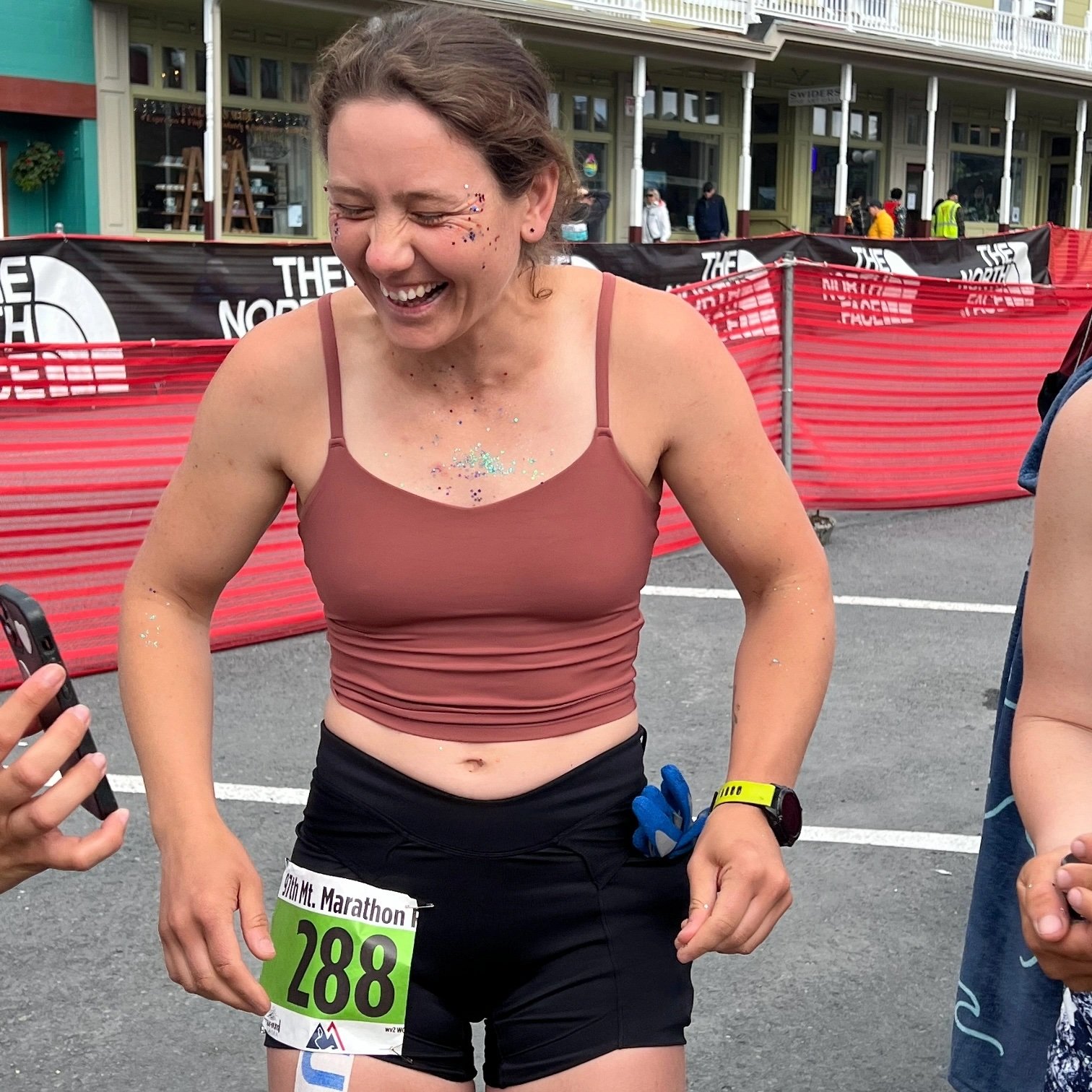 A woman with a broad smile, wearing a pink sports bra, black shorts, and a race bib numbered 288, standing outdoors after a marathon race, with glitter on her face and body, and people and red barriers in the background.