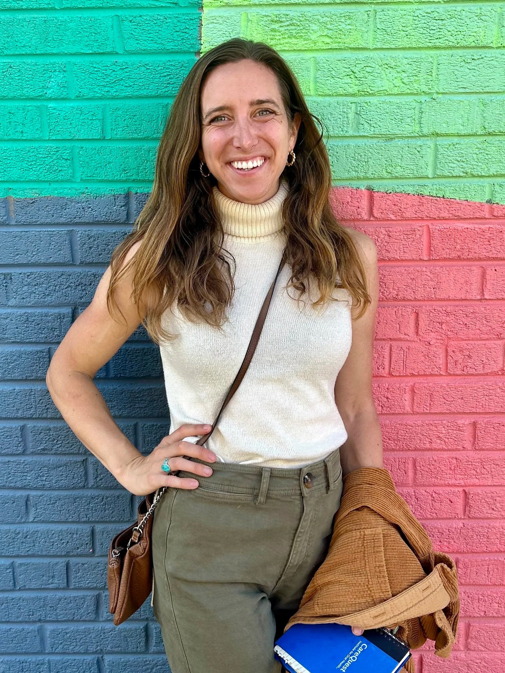A woman with long, wavy brown hair smiling while standing in front of a colorful brick wall painted with green, blue, and pink segments. She is wearing a sleeveless cream turtleneck top, olive green pants, and hoop earrings. She holds a brown jacket and a blue notebook in her left hand and has a small brown purse slung over her shoulder.